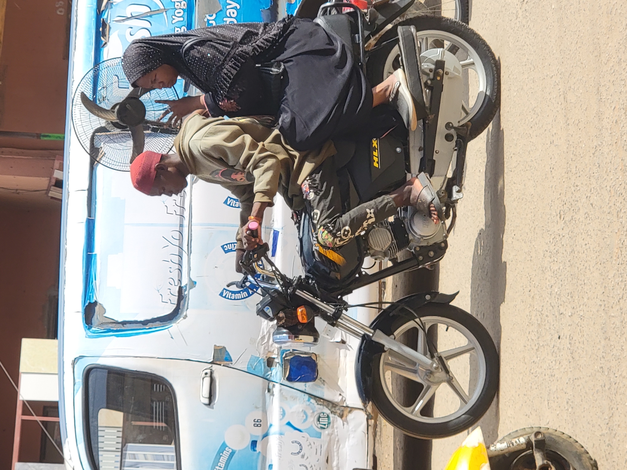 The image, taken in Maiburiji, Nigeria, captures a sunny outdoor scene featuring two individuals, a man and a woman, beside a motorcycle and a utility vehicle. The perspective is rotated 90 degrees clockwise, but when viewed upright, the subjects are interacting at ground level.
The man, wearing a red cap, a light brown long-sleeved shirt, and dark pants, is seated on a black motorcycle. His gaze is directed downwards and forward, and his hands are near the handlebars. The motorcycle has "HLX" prominently displayed on its side panel and features silver-colored wheels.
Beside him, a woman wearing a black hijab and a patterned black top leans forward. Her hands are positioned near a large, round standing fan with a metal cage, which is placed between her and the man. Her attention also seems to be directed forward, potentially at something the man is doing or an object in front of them.
In the background, a white and blue utility vehicle, possibly a delivery van or rickshaw, is parked. Its side features various branding and text, including "freshyo", "Vitamin", "Zinc", "tamins", and the numbers "98" and "08". Another tire or wheel is partially visible on the far right of the frame, lying on the light-colored ground or pavement.
The bright sunlight casts distinct shadows, suggesting it is mid-day or early afternoon. The presence of the fan indicates warm weather. The overall scene appears to be a casual street or market interaction.