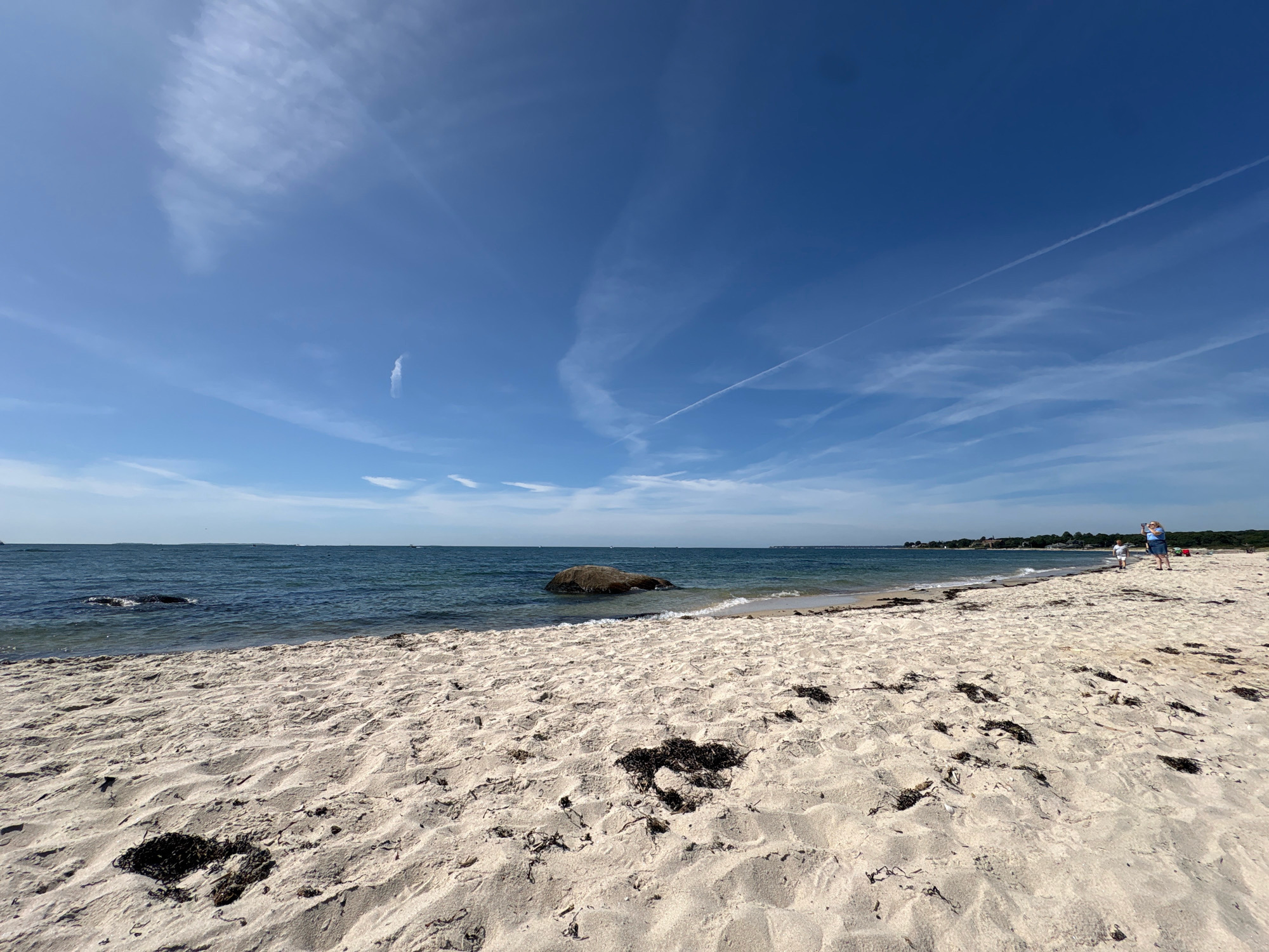 The image shows a beach scene in Waterford, United States of America. The foreground is dominated by a sandy beach with scattered dark seaweed. The middle ground features the ocean with a large rock partially submerged near the shore. The horizon is marked by a distant shoreline with trees and buildings. The sky is a clear blue with wispy white clouds and contrails from airplanes. In the far distance on the beach, two individuals are visible, one standing and taking a photograph. The overall scene depicts a sunny day at the coast.
