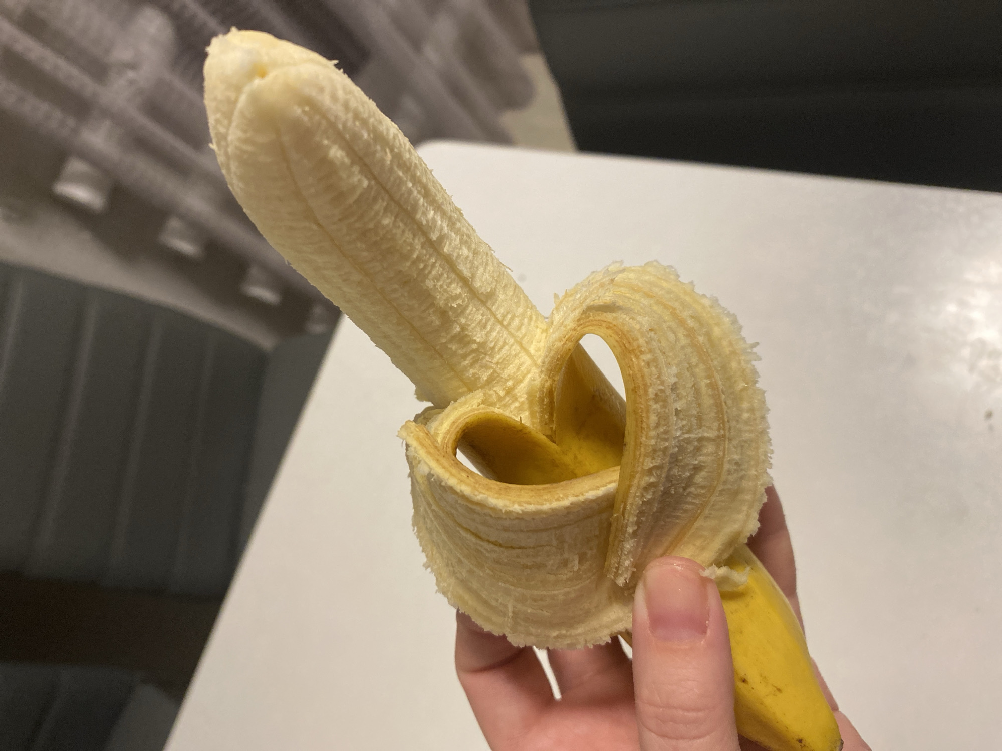 A close-up image shows a human hand holding a partially peeled, ripe banana indoors. The banana's yellow flesh is exposed, and its peel is folded back in such a way that it forms a distinct heart-like shape around the lower part of the fruit. The hand, with visible fingers and a thumb, supports the banana from beneath. The banana displays minor brown flecks on its unpeeled section, indicating ripeness.
The background is out of focus. A bright white surface, likely a table or countertop, occupies the lower and right parts of the background, while a darker, indistinct textured area, possibly a wall or blinds, is visible in the upper and left sections. The lighting suggests an indoor setting. The image captures a simple, everyday act of preparing to eat a banana, with the artful arrangement of the peel adding a notable detail. No specific time of day, weather conditions, or visible text can be determined from the image. The location context for this image is Noviy, Russia.