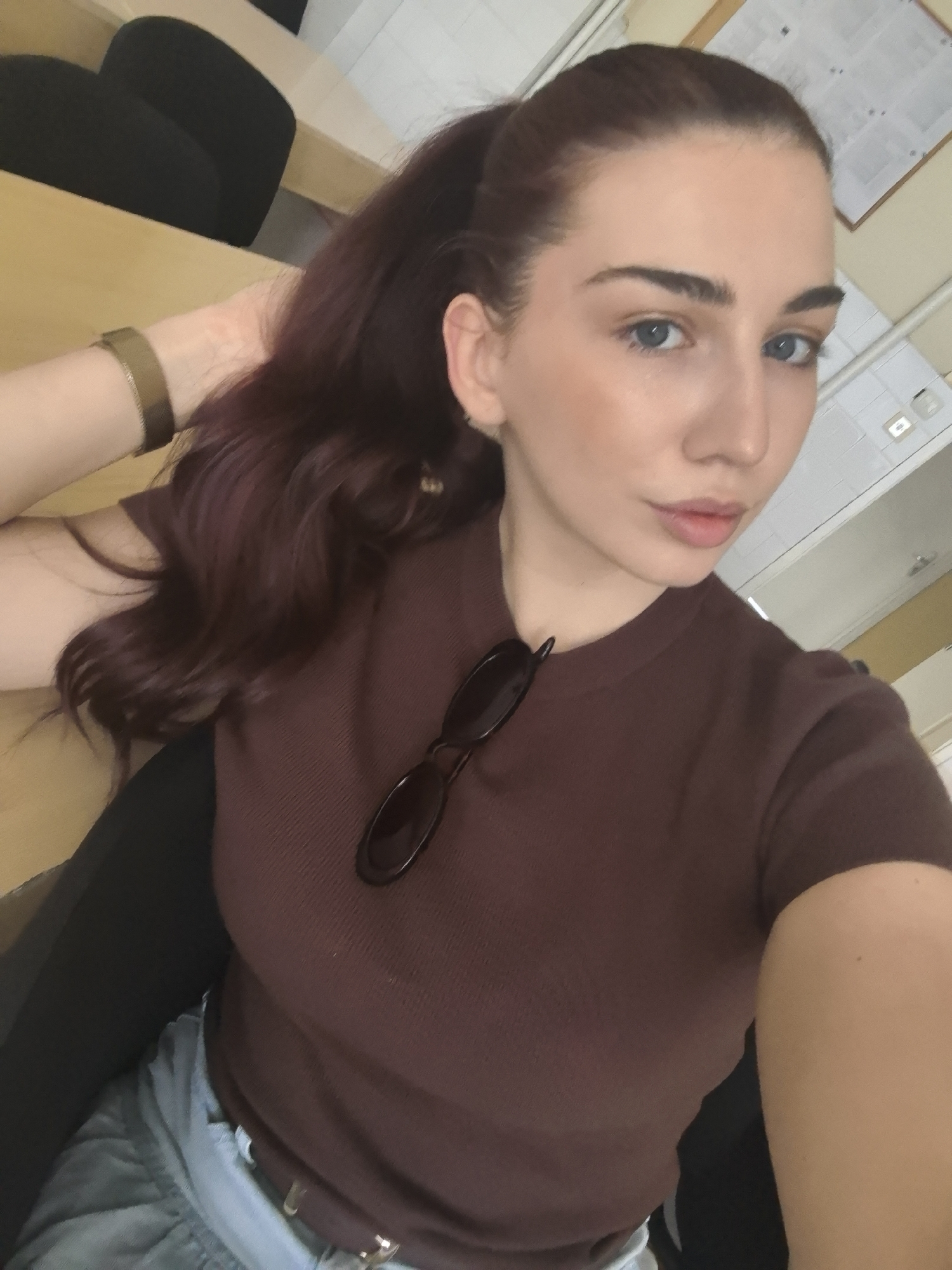 A young Caucasian woman with long, dark auburn hair pulled into a high ponytail is pictured in a selfie from the chest up. She is wearing a maroon, ribbed, short-sleeved shirt and light blue denim jeans. A pair of dark-rimmed sunglasses is resting on her chest, hanging from the neckline of her shirt. Her left arm is bent, with her hand resting behind her head, near her ear. She is wearing a thin, gold-colored bracelet on her left wrist. She has light blue eyes and appears to be wearing subtle makeup. The background suggests an indoor setting, possibly an office or classroom, with a wooden desk and black chairs visible. A bulletin board or framed document hangs on the wall in the upper right corner. The overall lighting is bright and natural, casting soft shadows. The image is shot from a slightly high angle.