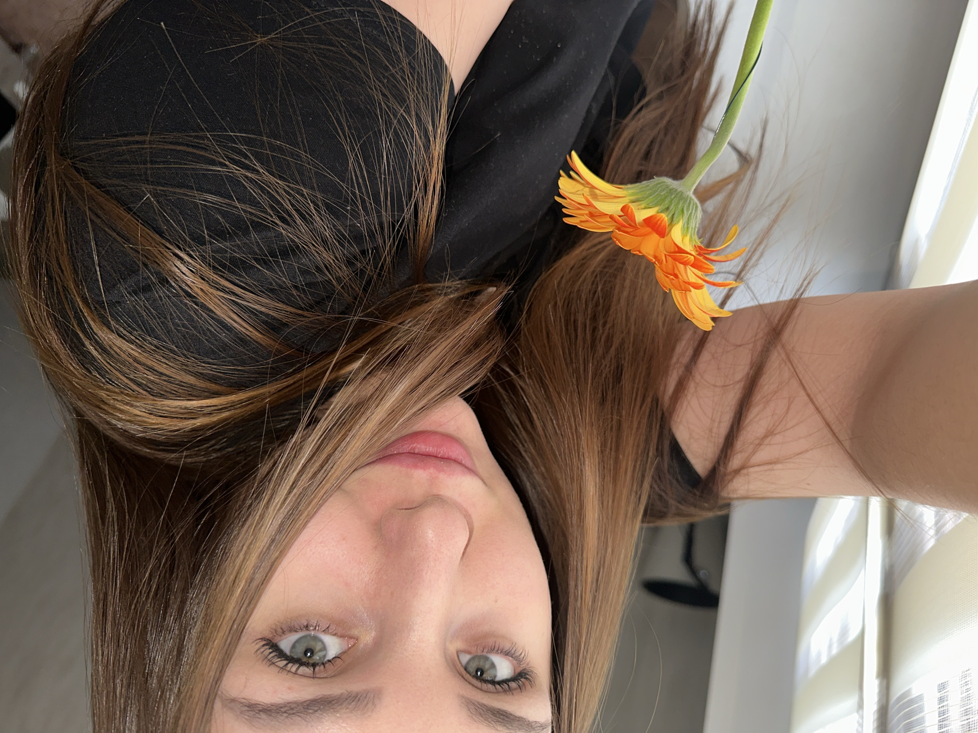 A close-up selfie shows a young woman with long, straight to wavy brown hair, featuring lighter highlights. She is looking upwards towards the camera with wide, light green or hazel eyes. Her lips appear natural with a hint of pink. She is wearing a black top or shirt, which frames her face and shoulder.
On the right side of the frame, she holds a vibrant orange and yellow gerbera daisy, its petals fanned out. The flower is fresh with a green stem, positioned near her hair and shoulder.
The setting appears to be indoors, likely a room. In the blurred background on the right, a window with white or off-white horizontal blinds is visible, indicating natural light source. The lighting is soft and even, highlighting her hair and complexion. The overall mood conveyed is calm and contemplative. No text is visible in the image. The location context provided is Chaikovsky, Russia.