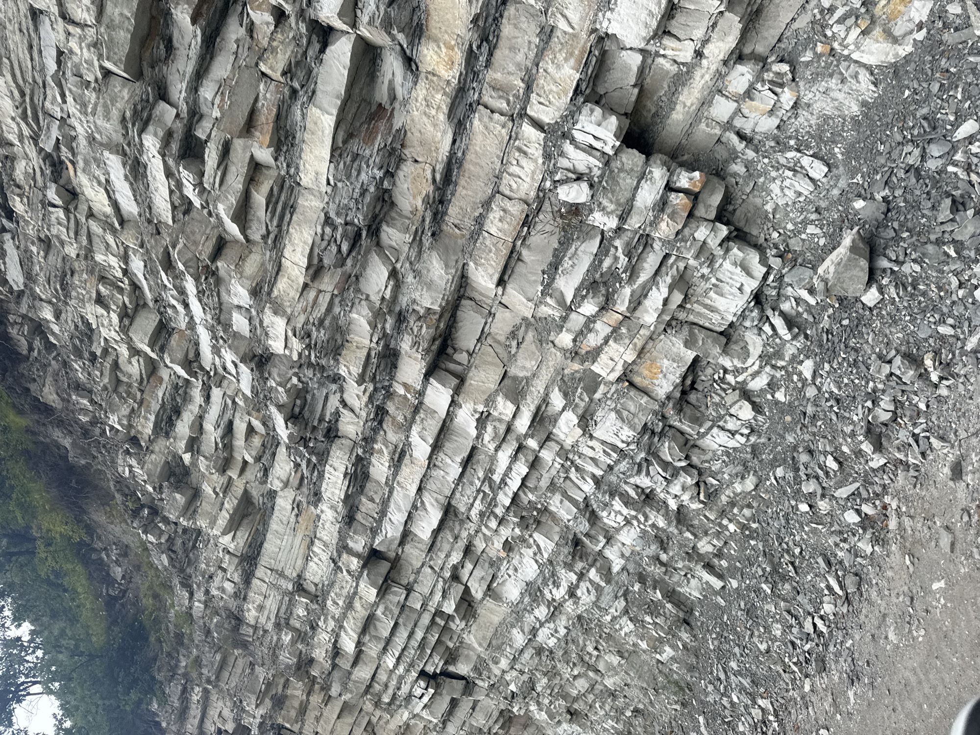This image captures a weathered rock face with distinct, horizontal striations, characteristic of sedimentary rock layers. The rock is light gray with darker gray bands interspersed, and some sections exhibit a yellowish-brown hue, suggesting mineral deposits. The rock face is steeply sloped and partially covered by a collection of loose, fragmented rocks at its base. A dirt and gravel path or road runs along the bottom left of the image. Above the rock face, a small area of green vegetation, including trees, is visible. There are no people or animals present in the scene. The overall impression is of a natural geological formation, possibly a roadside cut or a cliff face. The provided context indicates this scene is located in Nikhaloi, Russia.