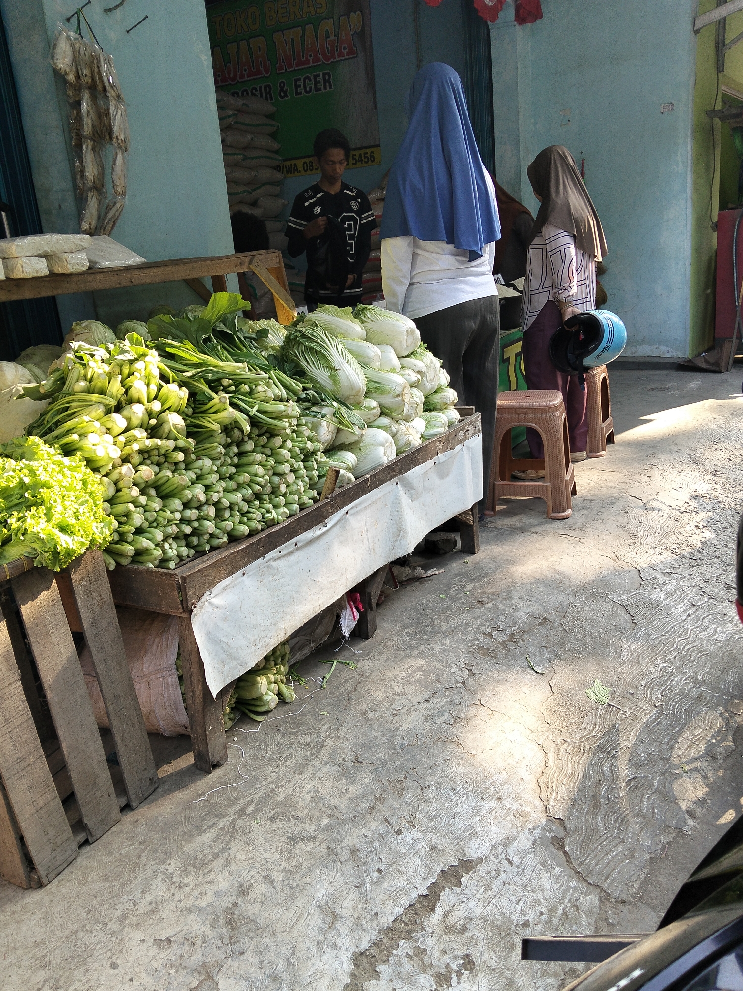 The image captures a bustling street scene in Karangmalang, Indonesia, likely outside a small market or shop. In the foreground, a wooden cart is laden with an abundant display of fresh vegetables, including lettuce, what appears to be bok choy or similar greens, and napa cabbage. The vegetables are neatly piled, suggesting they are for sale.
Behind the vegetable cart, a young man in a black t-shirt with white lettering is visible, seemingly engaged in tending to his stall. To his right, two women are standing. One woman is wearing a blue headscarf and a white top, and the other wears a brown headscarf and a patterned shirt with a maroon skirt. The woman in the brown headscarf is holding a light blue motorcycle helmet.
The backdrop of the scene features a building with a light blue facade, adorned with signs that read "TOKO BERAS" (Rice Shop) and "AJAR NIAGA" (Learn to Trade). Several large sacks, presumably filled with rice, are stacked behind the counter, further indicating the nature of the establishment. The ground is paved with concrete, showing signs of wear and tear, with sunlight casting dappled shadows across it. The overall impression is one of a typical, vibrant market environment in Indonesia.