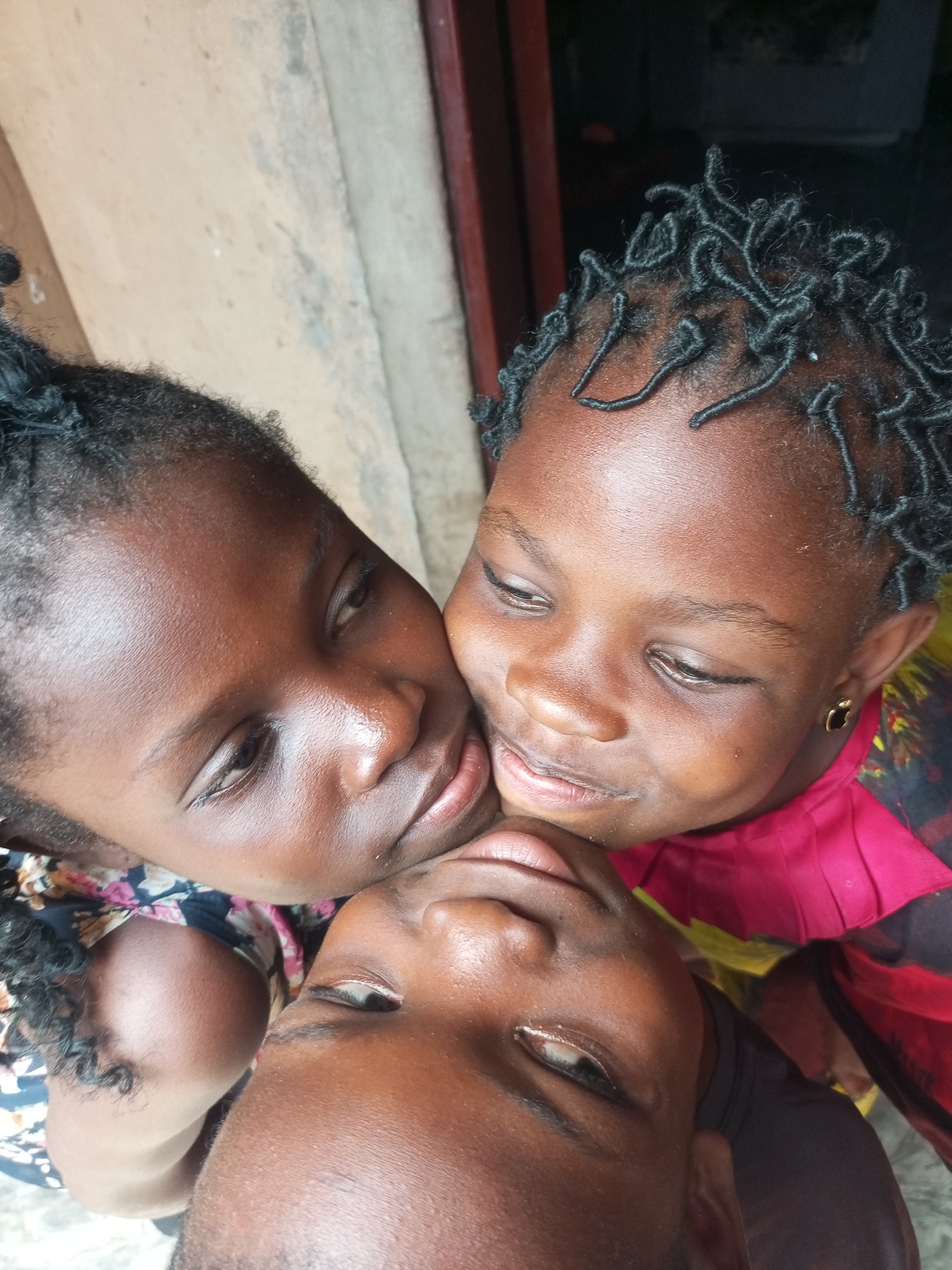 In Tempure, Nigeria, three individuals, two young girls and one person whose age and gender are indistinct, are captured in a close-up, overhead shot. The person in the foreground, whose face is partially visible, leans back, with their head tilted to the side. Two young girls are positioned on either side of this person. The girl on the left, with dark hair styled in intricate braids, is leaning in, her face close to the viewer's perspective. The girl on the right, also with braided hair and wearing a golden earring shaped like an apple, is similarly leaning in, their faces nearly touching. All three individuals have dark skin tones. The background is indistinct, but a textured wall and a doorway are partially visible, suggesting an outdoor or semi-outdoor setting. The lighting is natural, casting soft shadows.