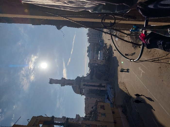 A landscape view shows a partially destroyed mosque in the foreground under a bright sun and cloudy sky. The surrounding area is war-torn with demolished buildings. A road cuts through the scene, populated by several individuals. An overhead view is partially obstructed by a balcony with hanging items and electrical wires. The scene conveys a sense of devastation and human presence amidst destruction.