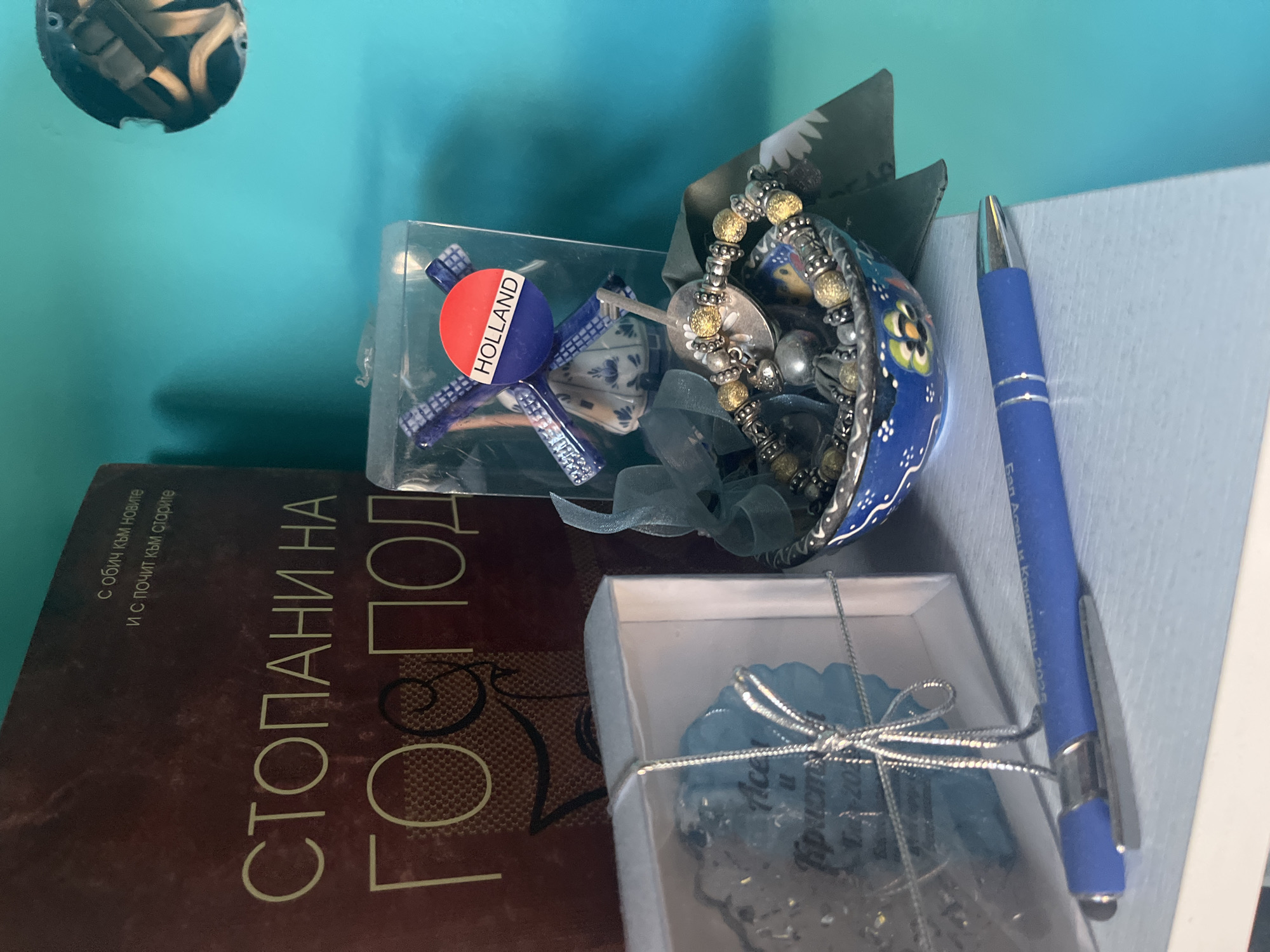 This image displays a still life arrangement of various objects on a light-colored surface, set against a vibrant teal-blue wall, located in the city of Pokrovnik, Bulgaria. On the left, a large, reddish-brown book or folder stands vertically, its cover bearing Cyrillic text; the prominent title reads "СТОПАНИ НА ГОСПОД" (Masters of the Lord), with a smaller inscription above stating "с обич към новите И с почит към старите" (with love for the new And with respect for the old). Adjacent to the book, a clear plastic box with a white base and a silver ribbon bow contains a translucent blue, flower-shaped decorative item, which has partially visible Cyrillic text, including "Асен и Кристиян" and a date fragment such as "18-202". In the central area, a small, dark blue ceramic bowl decorated with colorful floral motifs holds a bracelet composed of silver and gold-toned beads, along with a smooth, spherical silver charm and a small metallic, disc-like object. A sheer light-blue ribbon is draped over the items in the bowl. Behind the bowl, a green and white patterned piece of material is partially visible. To the right of the bowl, a miniature blue and white Delftware-style windmill ornament is encased in a clear plastic box, featuring a red circular sticker with "HOLLAND" in white text. A blue ballpoint pen, detailed with a silver clip and tip, lies horizontally on the foreground surface, displaying the inscription "Балкан Асен и Кристиян 2025." In the upper right portion of the image, a circular opening in the teal-blue wall exposes multiple light-colored electrical wires within an uncovered electrical junction box.