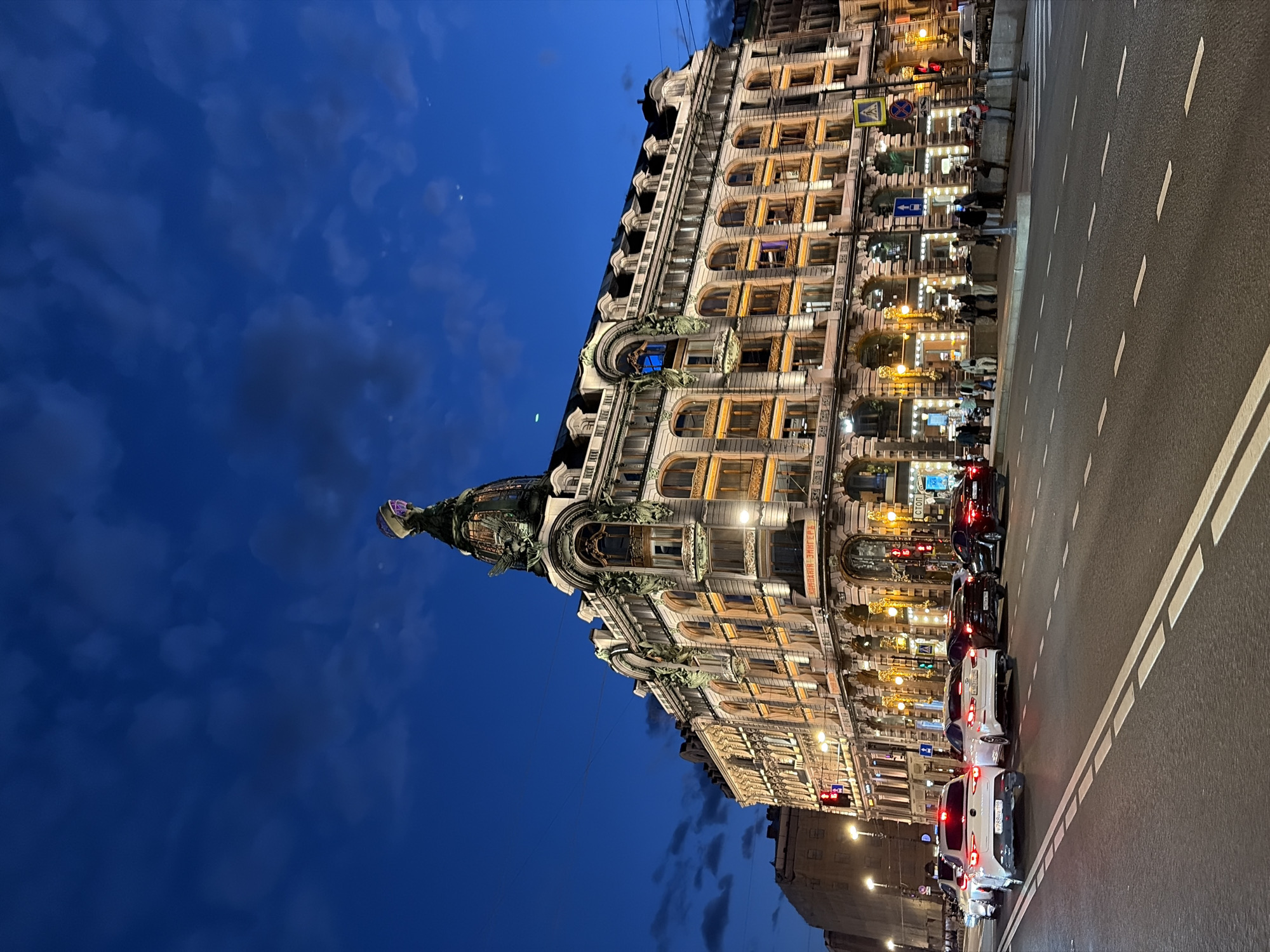 This is a nighttime, wide-angle, eye-level outdoor photograph of the Singer House (Dom Knigi) building in Saint Petersburg, Russia. The building is ornate and illuminated with warm yellow lights, highlighting its windows and architectural details. A distinctive glass dome topped with an eagle statue sits atop the building. The sky above is a deep blue, with scattered, wispy clouds. In the foreground, a paved road with white lane markings runs horizontally across the frame. Several cars are visible on the road, some with their headlights and taillights illuminated. People are walking on the sidewalk in front of the building, and streetlights are also visible. The overall scene depicts an urban streetscape at dusk or night.