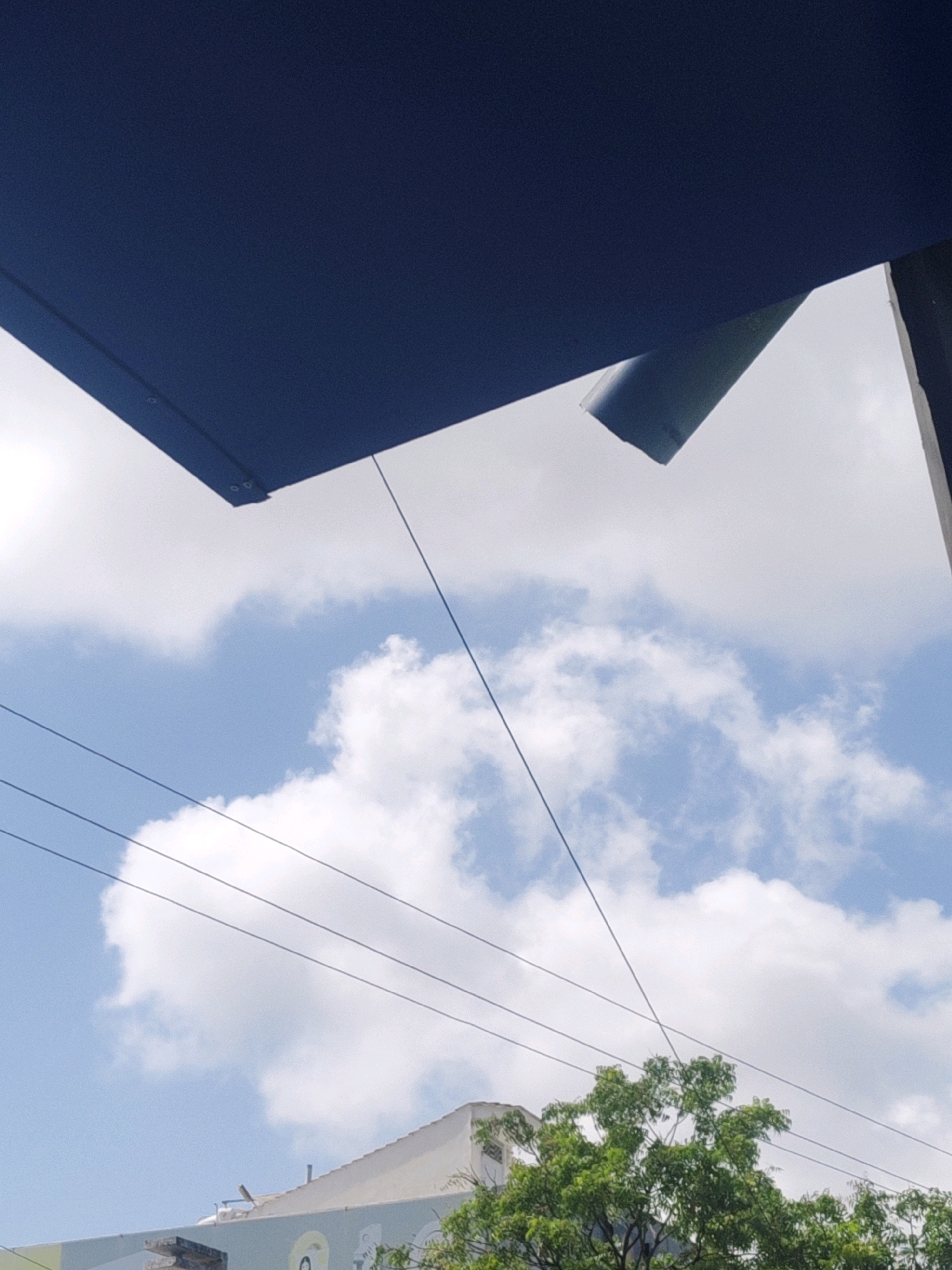 The image depicts an outdoor scene with a clear blue sky filled with white cumulus clouds. In the foreground, a large, dark blue structure, possibly a rooftop or awning, extends from the top left corner downwards. A portion of a metallic pipe or conduit protrudes from this structure. Several thin, dark wires are suspended across the sky, crisscrossing each other. Below the wires, the top of a building with a light-colored facade is visible, partially obscured by lush green foliage from a tree. The building appears to have some sort of mural or signage, with circular designs visible. The scene is indicative of an urban or semi-urban environment, possibly captured in Sumerpur, India, given the context. There are no people or overt actions depicted, focusing instead on architectural elements and the natural sky.