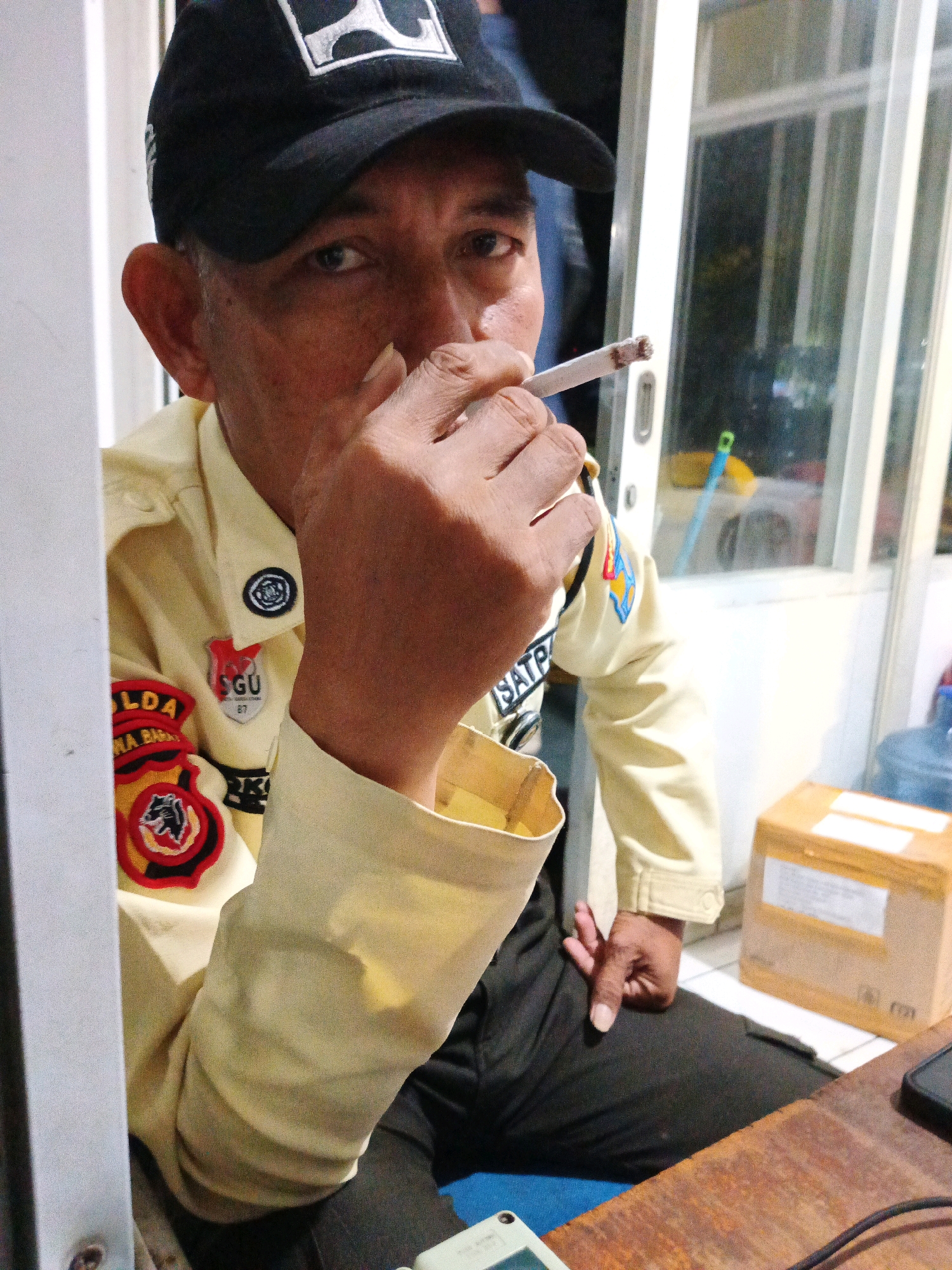 A man wearing a black baseball cap and a light-colored uniform is smoking a cigarette. The uniform has patches on the left chest and sleeve, one of which appears to read "LDA" and another with a crest. He is seated at a wooden desk, with a cardboard box and a water cooler visible in the background. The setting appears to be an indoor location, possibly an office or security post, with a window or glass door in the background. The image was taken in Bandung City, Indonesia.