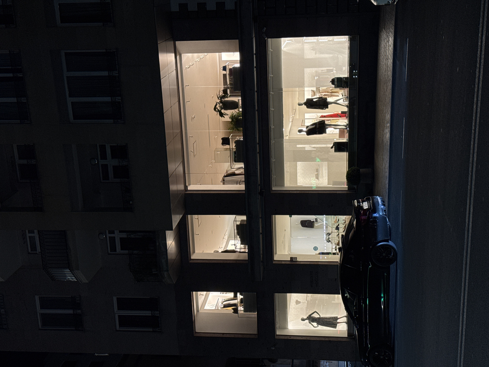 The image shows a multi-story building at night in Yerevan, Armenia. The building appears to be a combination of residential and commercial spaces. The residential part is visible on the left side with multiple windows, many of which have dark metal grates on the outside. The commercial aspect features large display windows showing mannequins dressed in clothing. A black car is parked in front of the building on the street. The street is paved and has white lines marking lanes. The overall lighting is low.