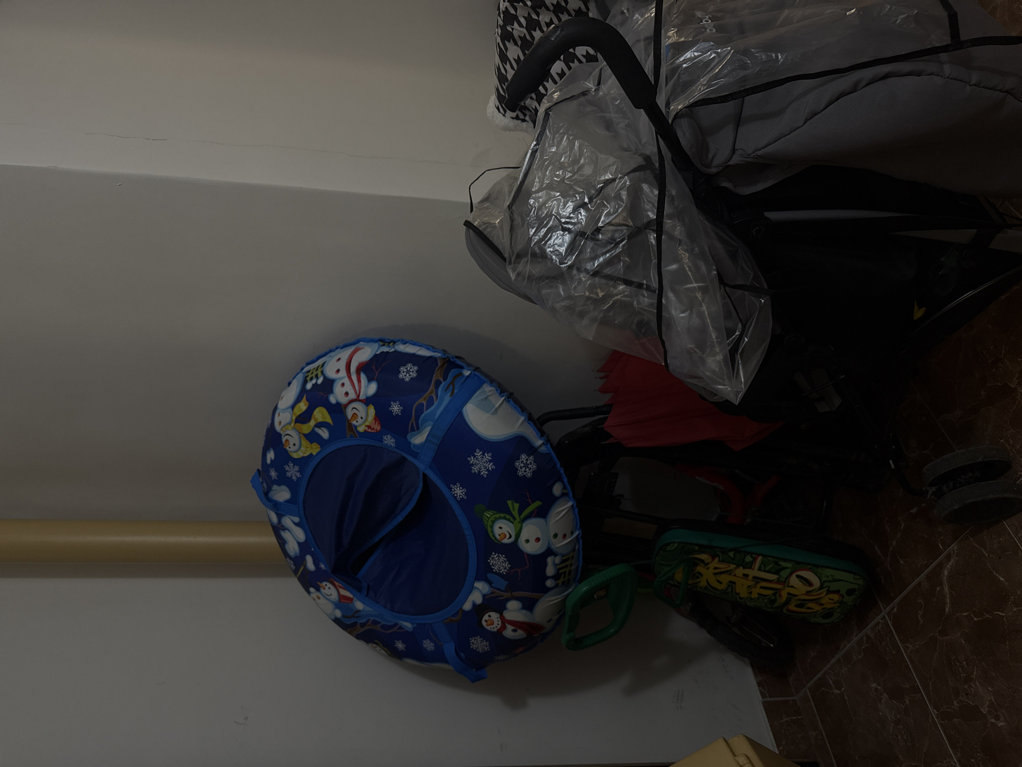 The image captures a dimly lit indoor scene, likely a storage area or a corner of a room, in Perm, Russia. The primary subjects are a baby stroller and a blue inflatable snow tube.
The stroller, positioned on the right side of the frame, is mostly covered by a clear plastic rain cover. A black and white houndstooth pattern fabric is visible on the upper part of the stroller, near its handle. The interior of the stroller, appearing dark gray, is visible through the plastic. Leaning against the wall is a vibrant blue inflatable snow tube, decorated with various cartoon snowmen and white snowflakes. Its central opening, also blue, is prominent.
Below the stroller and partly obscured, there is a dark green object with yellow, stylized, and illegible graffiti-style text. This object could be a skateboard or a similar wheeled toy. A piece of red fabric is also visible behind the snow tube, possibly part of the stroller or another item.
The walls are plain light gray or off-white. Several thin, hairline cracks are visible along the upper edge of the wall, where it meets the ceiling. A light brown, horizontal pipe or rod runs along the lower section of the wall. The floor consists of dark, possibly brown or reddish-brown, ceramic tiles. The lighting is low, creating a somewhat shadowed atmosphere. The word "baby" is faintly visible and upside down on the clear plastic cover of the stroller. No activity is depicted; the items appear to be stored.