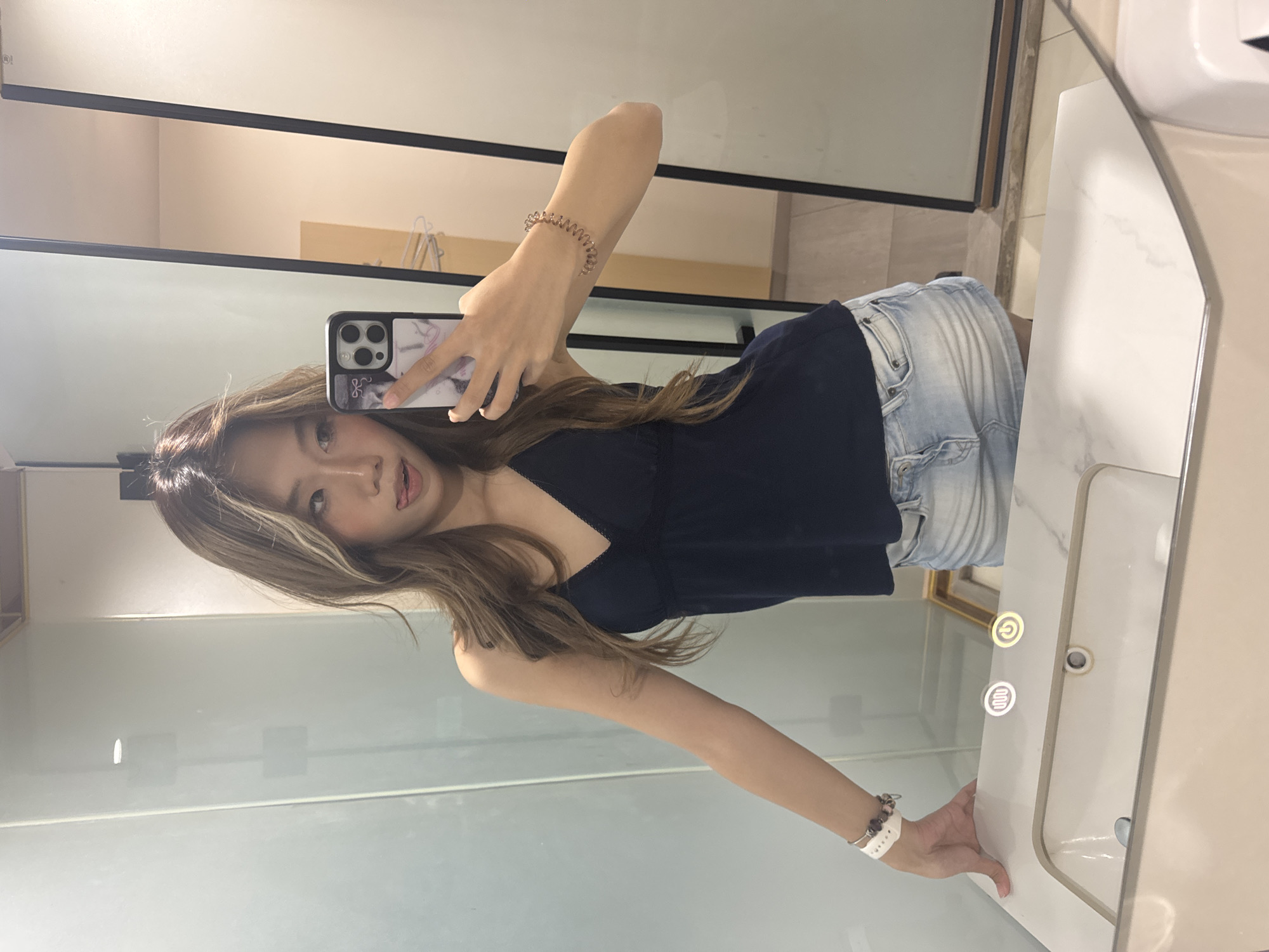 The image, taken in Yuzhong District, China, shows a young woman capturing a mirror selfie in an indoor setting, likely a modern bathroom or dressing room. The woman, with long, light brown hair featuring highlights, stands in front of a large mirror, angled slightly to her right. She holds a smartphone with a prominent multi-lens camera module and a decorative case in her right hand, extending her arm to take the photo of her reflection. Her expression is casual, with her mouth slightly ajar.
She is dressed in a dark blue or black camisole top with delicate lace trim and light blue denim shorts. Accessories include a clear, coiled bracelet on her right wrist and a white, perforated wristband on her left.
The background reveals a clean, contemporary space. A large mirror, framed in black, dominates the wall. To the right of the frame, a white vanity unit with a sink is visible. The vanity features two illuminated control icons: a circular power symbol and a symbol with four vertical bars, suggesting controls for lighting or other functions. The lighting in the room appears bright and even, indicating an indoor, well-lit environment.