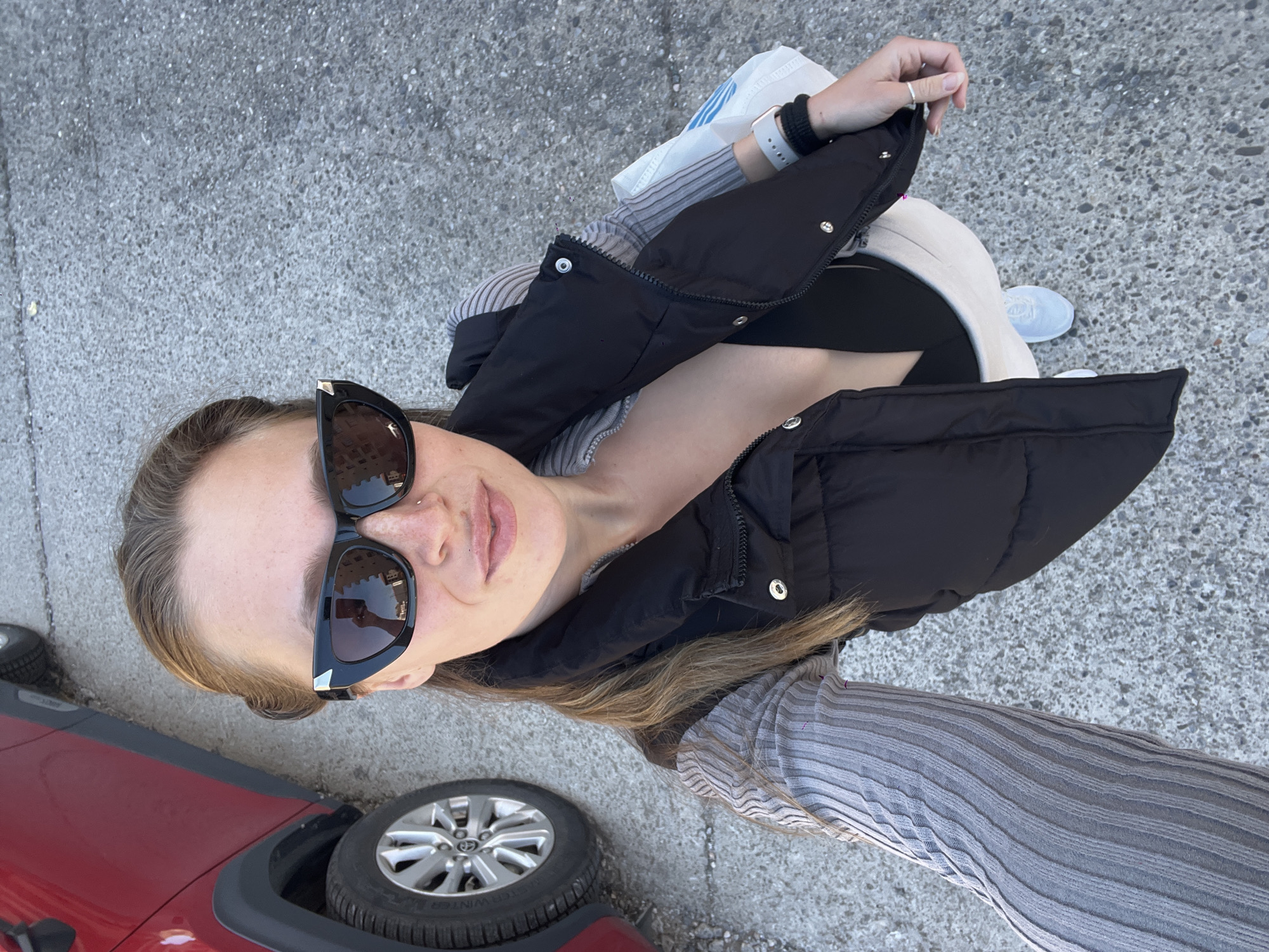 A young woman poses for a selfie on an overcast day next to a red car, possibly a Toyota based on the wheel emblem, parked on an asphalt surface. She is wearing dark sunglasses and a black puffer jacket over a striped top, with light-colored pants and white sneakers visible. A shopping bag is in her left hand. The image doesn't contain identifiable information to confirm the city.