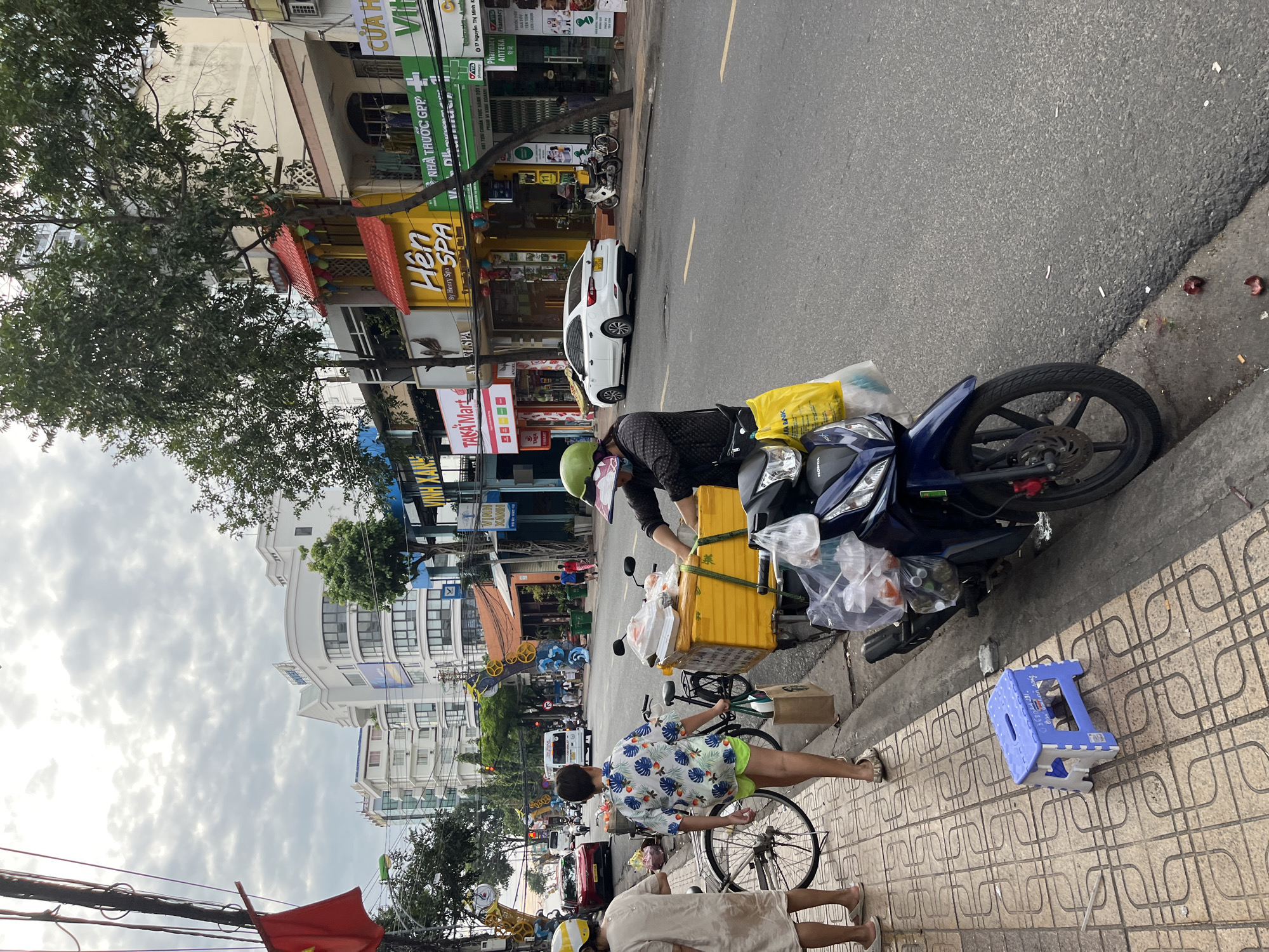 The image depicts a street scene in Nha Trang, Vietnam. In the foreground, a person wearing a green helmet and a dark patterned shirt is standing next to a blue motorcycle, which is loaded with a yellow container and several plastic bags of goods. The motorcycle is parked on the side of the road, next to a sidewalk paved with patterned tiles. A small blue plastic stool is on the sidewalk. To the left of the motorcycle, another person wearing a light-colored dress and a yellow helmet is walking away from the camera. Behind them, a bicycle is parked. The street is lined with buildings, including shops and businesses with colorful signage. Cars and other motorcycles are visible on the road. The sky is overcast.