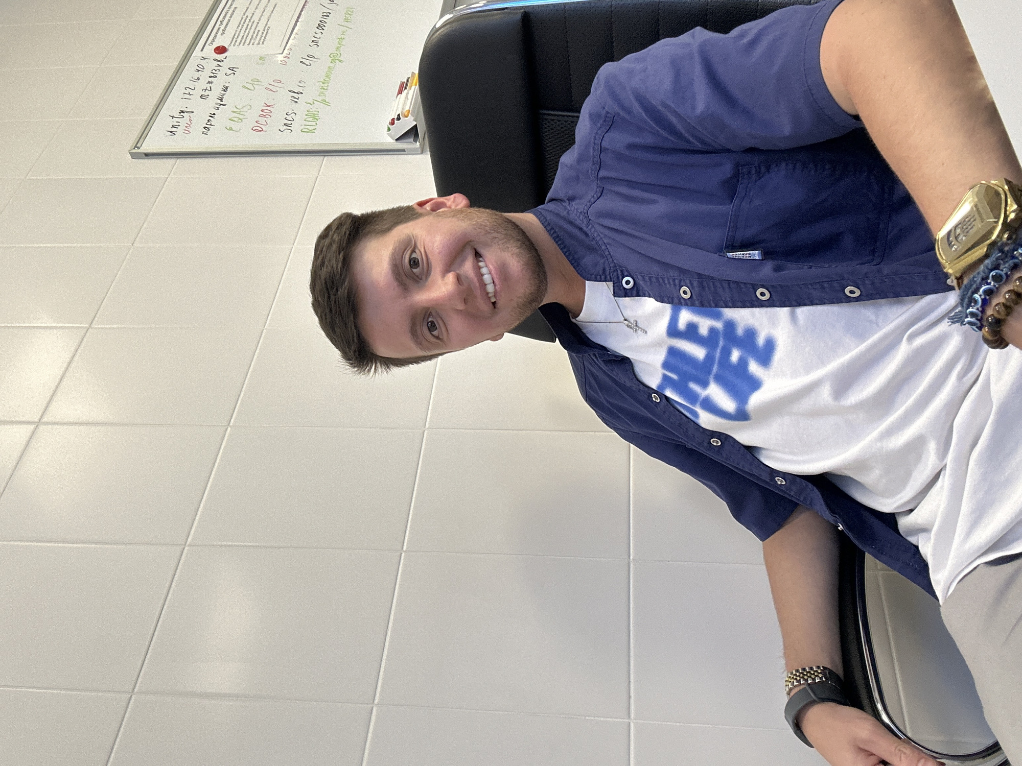 This is a portrait-oriented photograph of a young man sitting in an office chair. He is wearing a blue button-up shirt over a white t-shirt with blue lettering that appears to say "HLE LUFE". He has a watch on his left wrist, and a bracelet made of beads on his right wrist, along with another watch. The man is smiling and looking towards the camera.
The background consists of a white tiled wall and a whiteboard. The whiteboard has some handwritten text and diagrams on it, possibly related to IT or network information, as indicated by some visible text such as "Unity: 172.16.40.4" and "EQAS UP". The office setting suggests it might be daytime, but no specific weather information can be inferred. The overall impression is a casual and friendly snapshot taken in an office environment. The context of Moscow, Russia, is provided but not visually evident in the image itself.