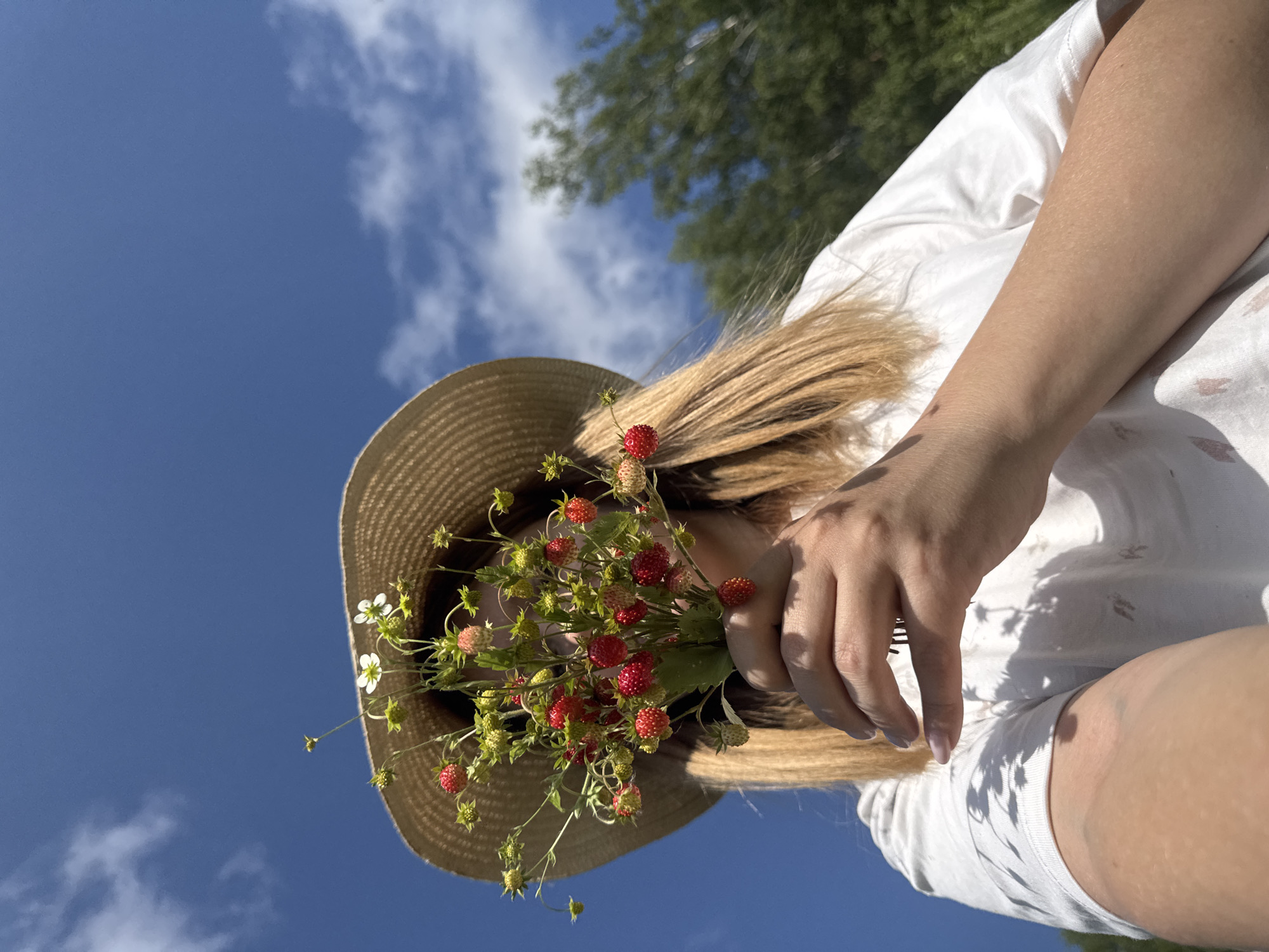 The image shows a person in Vassyata, Russia, holding a bouquet of wild strawberries and small white flowers. The person is wearing a straw hat and a white t-shirt, and their face is obscured by the bouquet. The background is a bright blue sky with some scattered clouds, and green trees are visible in the distance. The lighting suggests it is daytime, likely sunny. The perspective is from below, looking up at the person.