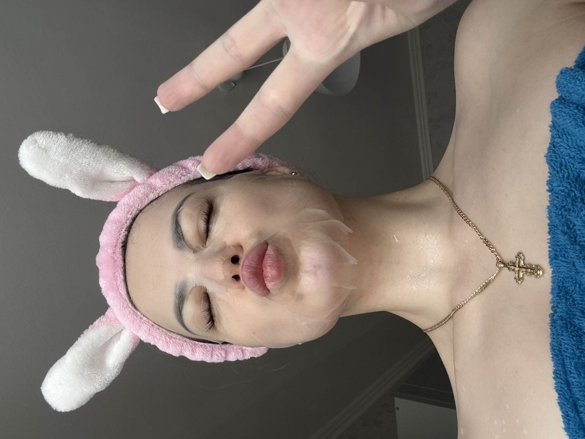 A woman wearing a pink bunny-ear headband and a transparent face mask is shown with her eyes closed and lips pursed. She is making a peace sign with one hand. A gold cross necklace is around her neck, and a blue towel is draped over her shoulder. The background is a neutral grey. The location is Unknown, Unknown.