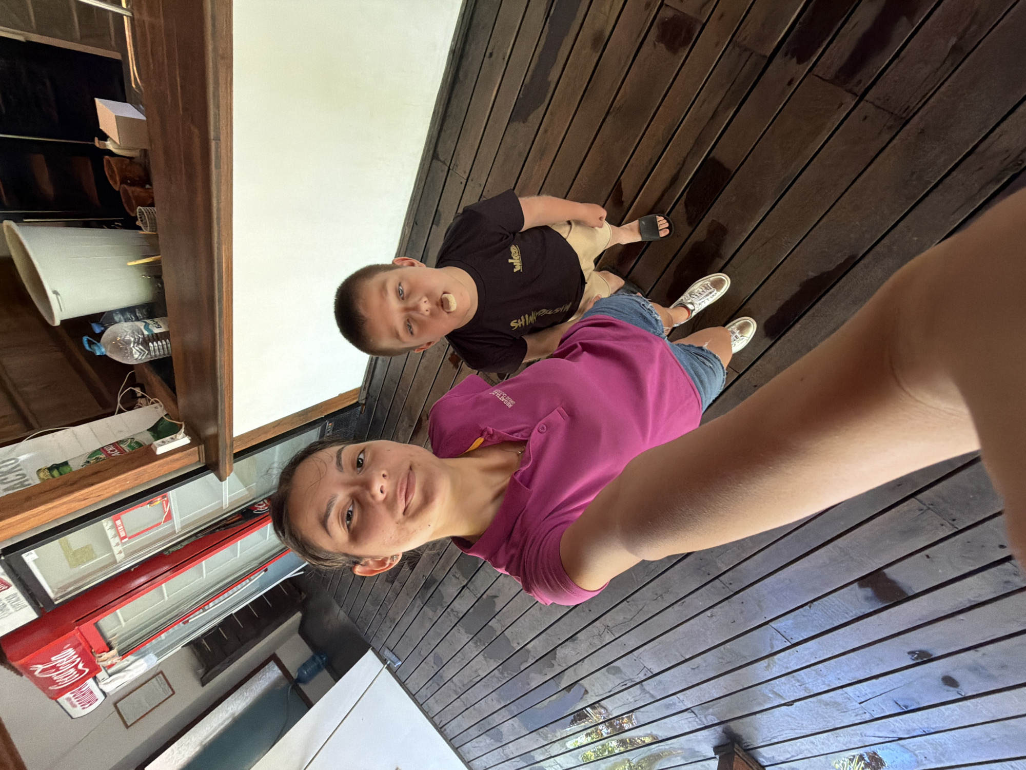 This media file captures a candid moment of two people on a wooden deck. The subjects are a young boy and a young woman. The boy, positioned in the upper half of the frame, wears a black t-shirt and tan shorts, with a small object, possibly food, in his mouth. He is looking directly at the camera with a playful expression. The young woman is in the foreground, holding the camera to take a selfie. She wears a bright pink t-shirt and denim shorts. Her gaze is also directed towards the camera, and she appears relaxed and happy.
The setting is an outdoor wooden deck, with planks visible throughout the image. The deck appears to be slightly wet, suggesting recent rain or a damp environment. In the background, to the left, there are glimpses of what seems to be an interior space with shelves holding various objects, including bottles and what looks like a white container or bin. There is also a red Coca-Cola branded cooler or refrigerator visible.
The lighting suggests it is daytime, possibly late morning or afternoon, with natural light illuminating the scene. The overall mood is casual and lighthearted, indicative of a relaxed outing or vacation. The image is oriented sideways, characteristic of a selfie taken with a phone. No explicit text indicating the time of day or specific event is clearly legible, though the Coca-Cola branding offers a hint of the commercial environment. The location context provided is Tsarevo, Bulgaria.
