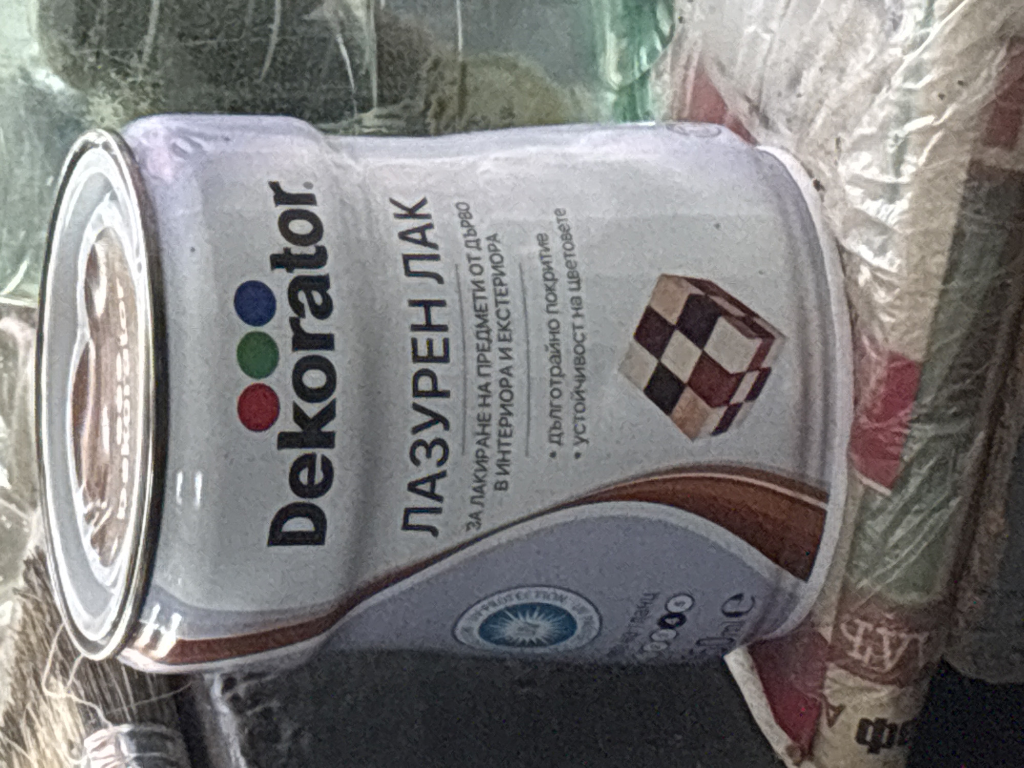 The image shows a close-up of a can of "Dekorator" brand varnish. The can is primarily white with red, green, and blue dots above the brand name. Below the brand name, it reads "ЛАЗУРЕН ЛАК" (azure varnish) in larger text. Further text on the can describes it as being for varnishing wooden objects for indoor and outdoor use, with a promise of long-lasting coverage and colorfastness. A small checkered tile pattern, resembling wood blocks, is visible on the lower part of the can. The can is sitting on a pile of what appears to be woven bags, possibly containing rice or similar granular material. The background is out of focus but suggests a cluttered environment, possibly a market stall or storage area. The overall lighting is somewhat dim, with a slight warm cast. There is no indication of specific location within Plovdiv, Bulgaria, beyond what can be inferred from the Cyrillic script on the product label.