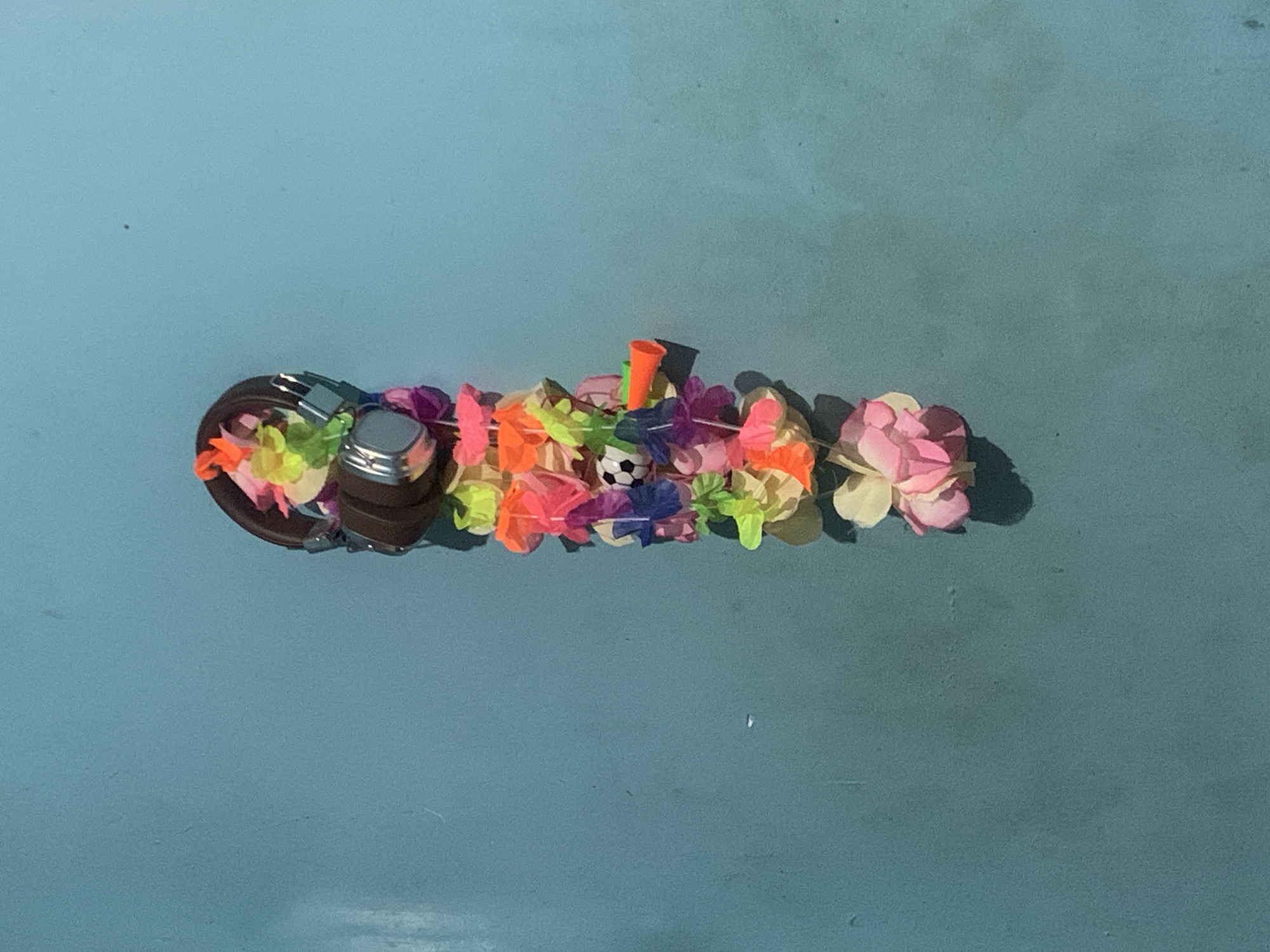 This close-up image features a decorative arrangement set against a uniform, light blue background. The main subject is an elongated cluster of vibrant, artificial flowers, appearing to be made of paper or fabric, in various colors such as pink, orange, yellow, green, and purple. These flowers are loosely arranged, resembling a miniature garland or lei.
Nestled within the center of this floral display is a small, white and black soccer ball. To the right of the soccer ball, a small orange cone-shaped object and a thin green stick are partially visible, peeking out from among the flowers. To the left of the floral arrangement, a brown-strastrapped object, possibly a watch or bracelet with a silver-toned metal buckle and case, is visible. The objects cast soft shadows, indicating a light source from the upper left.
The background is a consistent, plain light blue surface, which could be a table, wall, or floor. There are no people present, and no discernible activity is occurring; the items are presented as a still-life display. The lighting is even, suggesting an indoor setting or diffuse outdoor light. No text is visible in the image. The scene is located in Santa Ana, Venezuela.