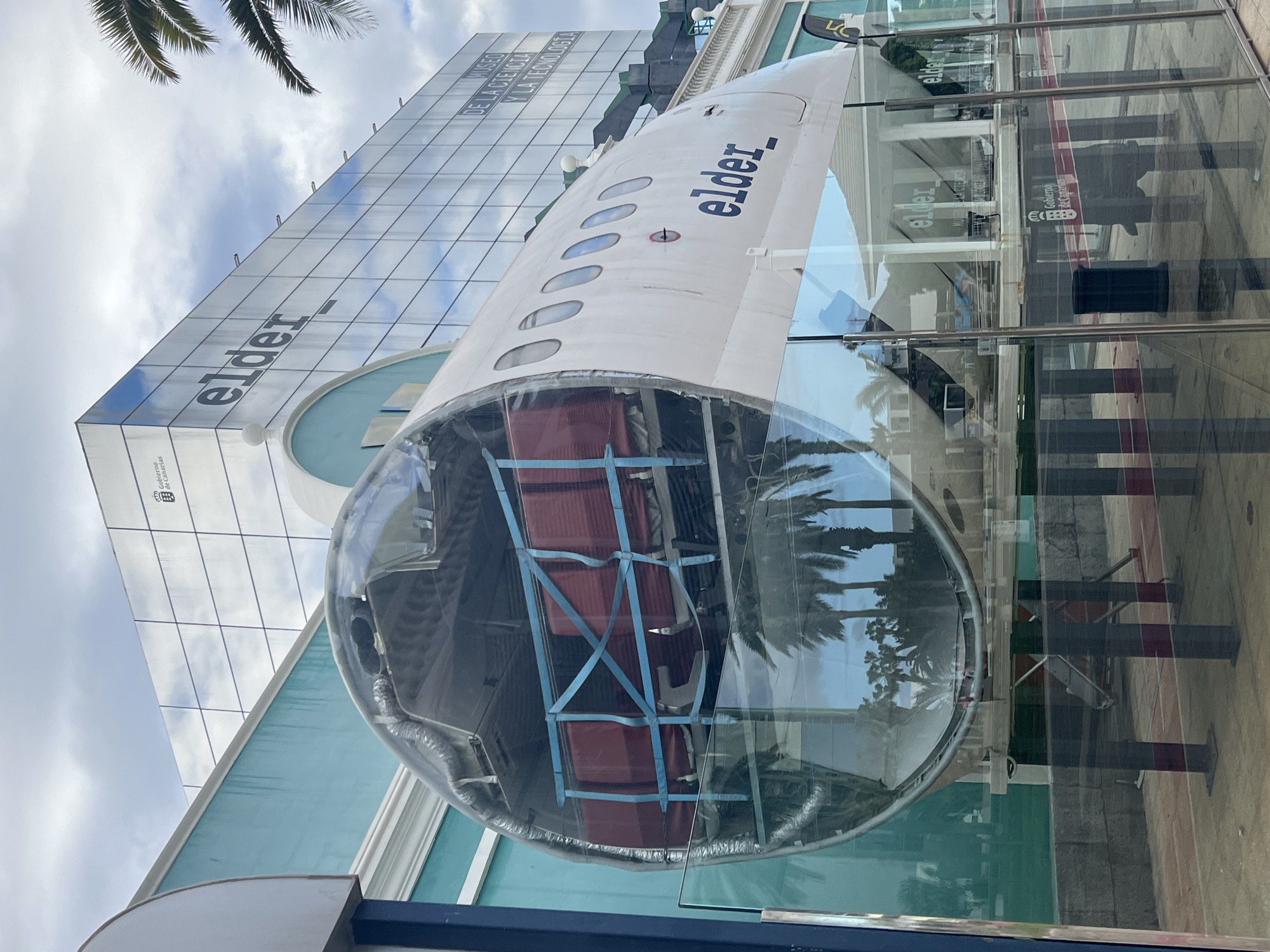 The image captures an outdoor display featuring a section of an airplane fuselage positioned in front of a modern glass-fronted building, all rotated 90 degrees clockwise. The scene is illuminated by daylight under a partly cloudy sky, with palm fronds visible in the upper left corner and reflected in the glass surfaces.
The prominent subject is a large section of a white airplane, marked with the black text "elder_" on its side, visible against the reflections of the sky and the building. The front part of this fuselage section is transparent, revealing an interior structure with red components, possibly seats or cargo, secured by blue straps or netting. This airplane segment is angled, with its transparent end facing the viewer.
Behind the airplane is a tall, contemporary building with an extensive glass facade. The word "elder" is boldly displayed in large black letters on the building's exterior. Below this, further text is visible: "MUSEO DE LA CIENCIA Y LA TECNOLOGIA" (Museum of Science and Technology), along with a logo and "Gobierno de Canarias" (Government of the Canary Islands). The glass panels of the building reflect the sky, clouds, and surrounding elements, including other parts of the building and palm trees.
The setting suggests a public display, likely outside the Elder Museum of Science and Technology in Las Palmas, Spain, given the explicit text. No people or active events are depicted; the image focuses on the static architectural and aeronautical elements. The overall impression is one of modern architecture and scientific exhibition.