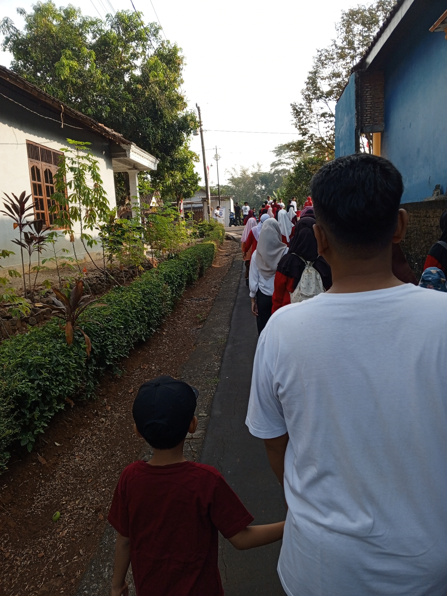 An outdoor, eye-level, medium shot captures a group of people walking in single file down a narrow, paved path. The path is bordered on the left by a white, single-story building with a wooden-framed window and lush green plants, including a row of manicured bushes. A brown-tiled roof extends over the building's eaves. To the right of the path, a tall, blue, two-story building with a brick facade is visible. The sky above is bright and overcast.
In the foreground, a young boy wearing a red t-shirt and a black baseball cap is walking to the left, holding hands with an adult male in a white t-shirt. The adult's back is to the camera, and the boy's back is also to the camera.
Further down the path, the group comprises at least ten individuals, mostly women and girls, many wearing headscarves and brightly colored clothing, including red, white, and grey. One person in the middle of the group is carrying a light-colored backpack. Some trees with green foliage are visible in the background. The ground beside the path is covered in dirt and small pebbles, with some scattered leaves. A single green object, possibly a fruit, lies on the dirt to the left of the path. The scene appears to be in a residential area in Lemahbang, Indonesia, given the architectural style of the buildings and the vegetation.