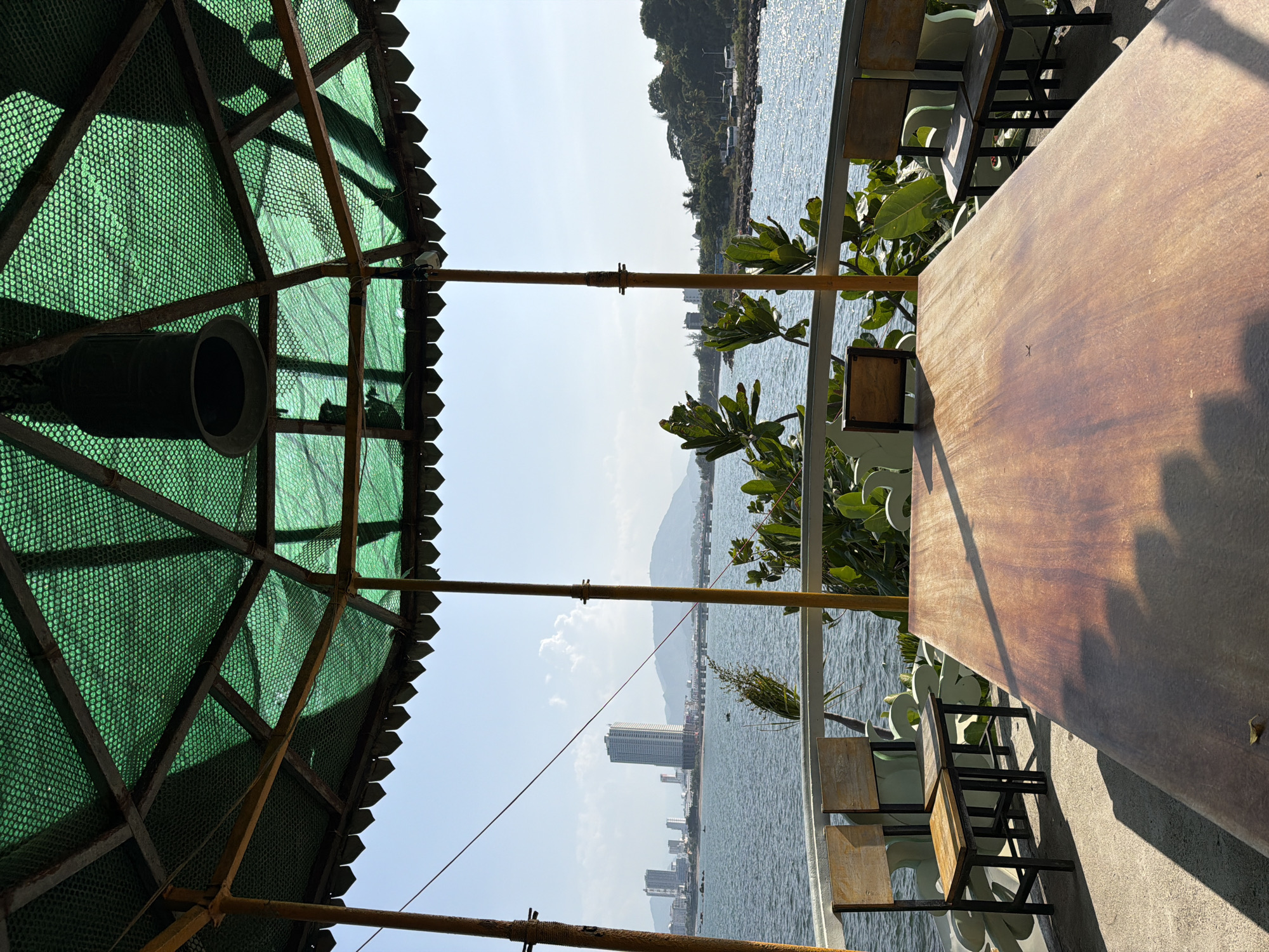 This is a low-angle shot of a wooden table and chairs under a green, netted canopy structure, looking out at a body of water. In the distance, a city skyline with several tall buildings, a bridge, and mountains can be seen. The water is calm and reflects the clear blue sky. The scene appears to be an outdoor seating area, possibly a restaurant or cafe, overlooking the coast of Nha Trang, Vietnam. The architecture of the canopy suggests a tropical or coastal setting. The lighting indicates it is daytime, with sunlight casting shadows on the table and ground. No people are visible in the frame.