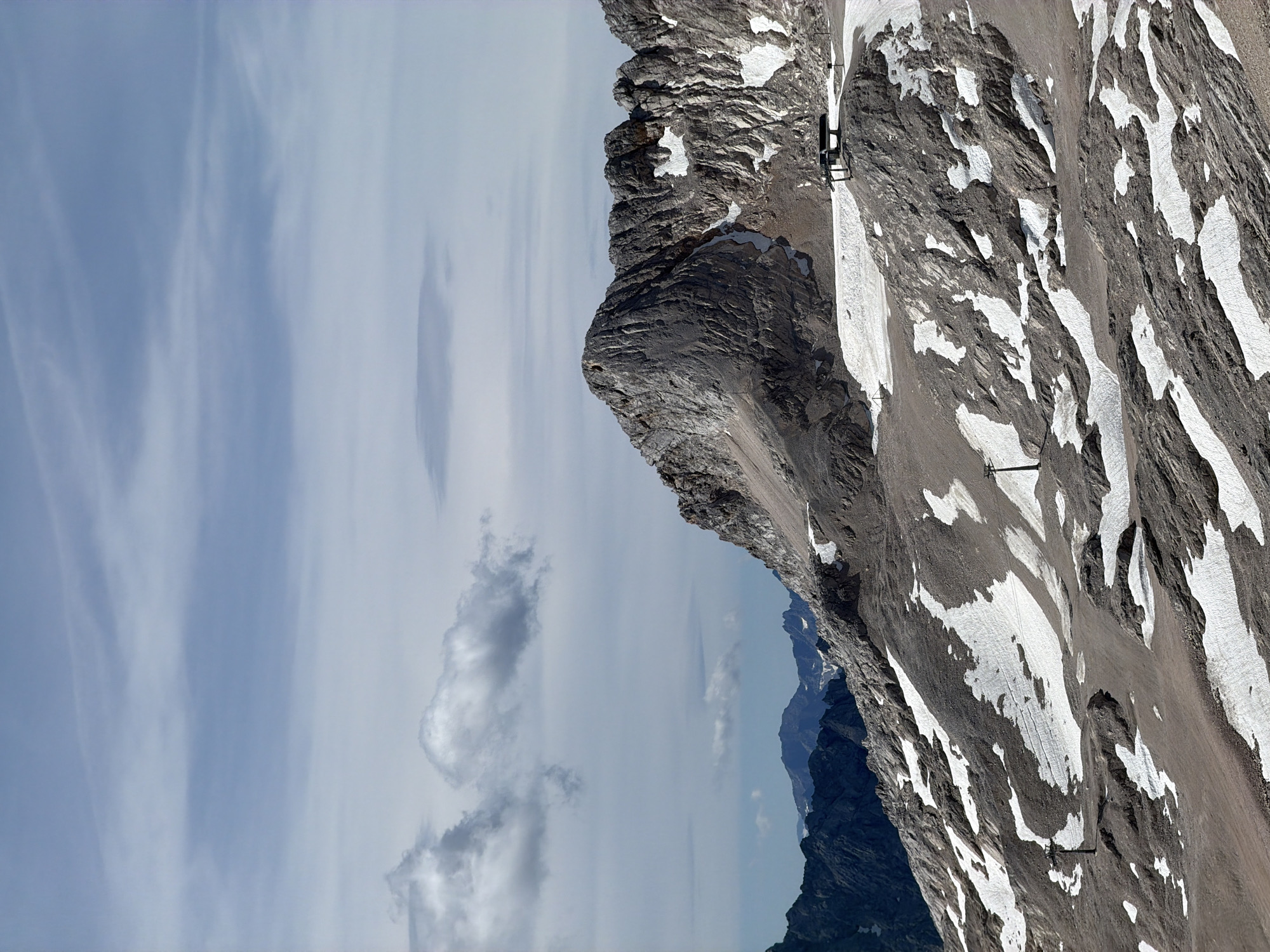 The image depicts a mountainous landscape in the Zugspitze region near Garmisch-Partenkirchen, Germany. The foreground features a rocky, barren slope with patches of snow scattered across its surface. Several ski lifts, identifiable by their poles and cable systems, are visible on the slope. One ski lift station, a small structure with a roof, is situated on a higher rocky outcrop. In the background, a large, jagged mountain peak dominates the scene, with its rocky slopes also showing signs of snow. Further in the distance, a range of darker, more densely packed mountains is visible under a cloudy sky. The sky itself is a pale blue with scattered white and grey clouds. The overall impression is one of a high-altitude, alpine environment, likely during a time when snow is present but not covering the entire landscape.