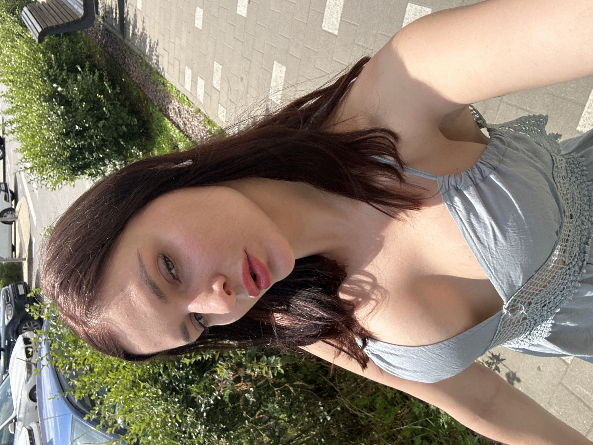 A young woman with long, dark brown hair and light skin is taking a selfie outdoors. She is wearing a light blue dress with thin straps and a crocheted accent. Her lips are painted with a pinkish-red lipstick. The background shows a paved walkway with a brick pattern, green bushes, and parked cars, suggesting an urban or suburban setting. Sunlight is casting shadows, indicating it is daytime.