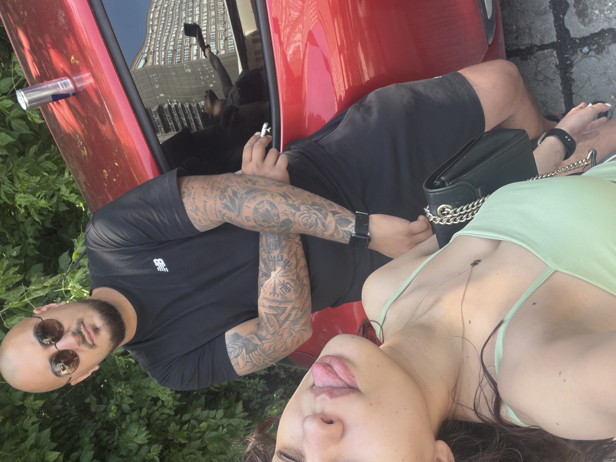 The image shows two people posing outdoors, appearing to lie horizontally across the frame due to the photo's orientation.
In the foreground, a woman with long dark hair is visible, wearing a light green strap top and a delicate gold chain necklace. She has a playful expression, with her tongue sticking out. A black wallet or clutch bag with a gold chain strap rests near her.
Behind her is a man with a bald head, a beard, and round sunglasses. He wears a black t-shirt with a white "NB" (New Balance) logo and black shorts. Both his arms are heavily tattooed with intricate designs, including clock faces, geometric patterns, and some partially visible text, one line of which appears to be "...we grow in different directions yet remain of one...". He holds a lit cigarette between his fingers and wears a black smartwatch on his left wrist.
The man leans against a red car, which features a silver Red Bull can (with "Red Bull" text and barcode visible) wedged in its partially open window. The car's reflection clearly shows a tall, modern building with many windows, a blue sky, and a person holding a phone, likely taking the photo. The background includes lush green foliage and visible cracked pavement, suggesting an urban or suburban outdoor setting during daytime. The overall atmosphere is casual and relaxed.