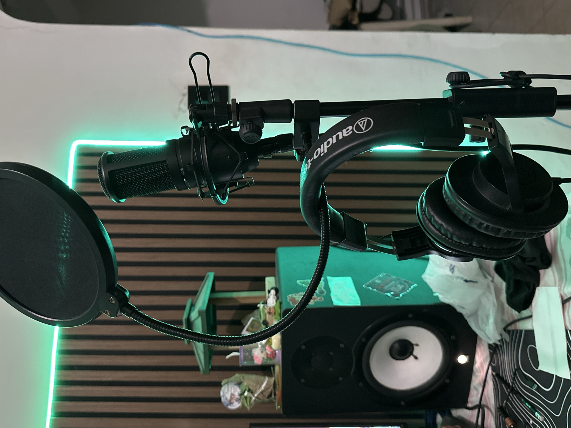 The image captures an indoor studio setup, potentially within a residence in Sabana de Guacuco, Venezuela. The scene is illuminated by a vivid green LED light strip installed horizontally along the top edge of a wooden slatted wall and continuing vertically down the right side.
At the center-right, a black condenser microphone, enclosed in a shock mount, is affixed to a black vertical microphone stand. A black circular pop filter is positioned in front of the microphone via a flexible gooseneck arm. Directly below the microphone on the stand, black over-ear Audio-Technica headphones are suspended, with the brand logo prominently displayed on the headband, reflecting the green light.
To the left of the microphone stand, a black studio monitor speaker is present, featuring a large white woofer cone and a smaller black tweeter. The top surface of this speaker serves as a display area for various small decorative items, including a green pagoda-like structure, a clear cup containing colorful miniature objects, a small figurine, and a white toy car. The side of the speaker has several stickers applied to it, some glowing with the reflected green light.
The background behind the speaker consists of a wall clad with vertical wooden slats or acoustic panels. To the right, a plain white wall or architectural element is visible, with a thin blue cable running vertically along its surface. The foreground reveals a patterned black and white mat, on which a small brass-colored object and a white rectangular piece of paper are located. A crumpled white plastic bag and a piece of dark fabric are also present on the surface near the speaker. No individuals are depicted in the image.