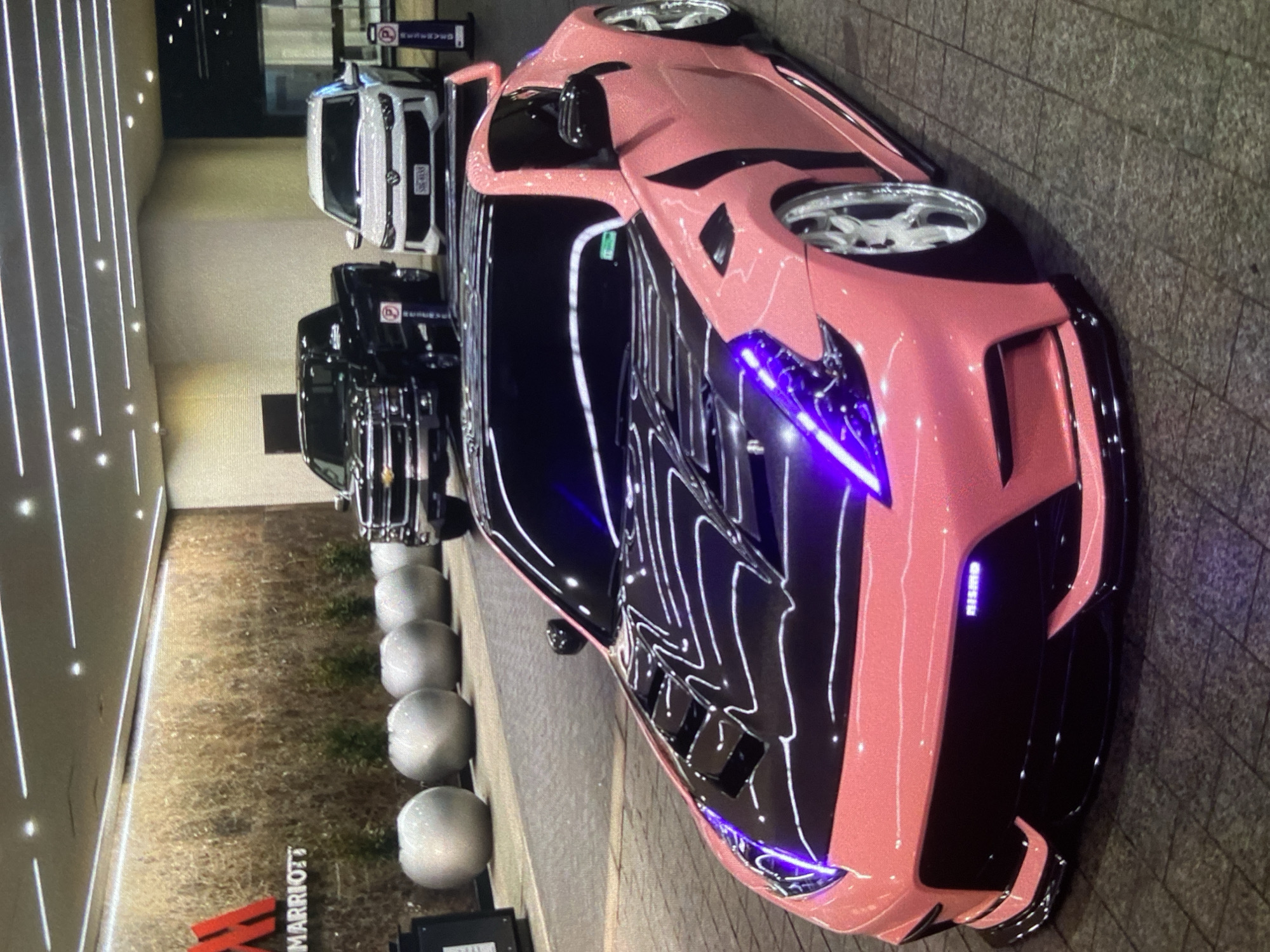 The image shows a pink and black modified Nissan sports car parked in front of a Marriott hotel. Several other vehicles are parked behind it. The cars are parked on a paved surface. The hotel is visible in the background. It is impossible to determine if the image was taken in Lhokseumawe, Indonesia. There is no indication of people, interactions or actions in the photo.