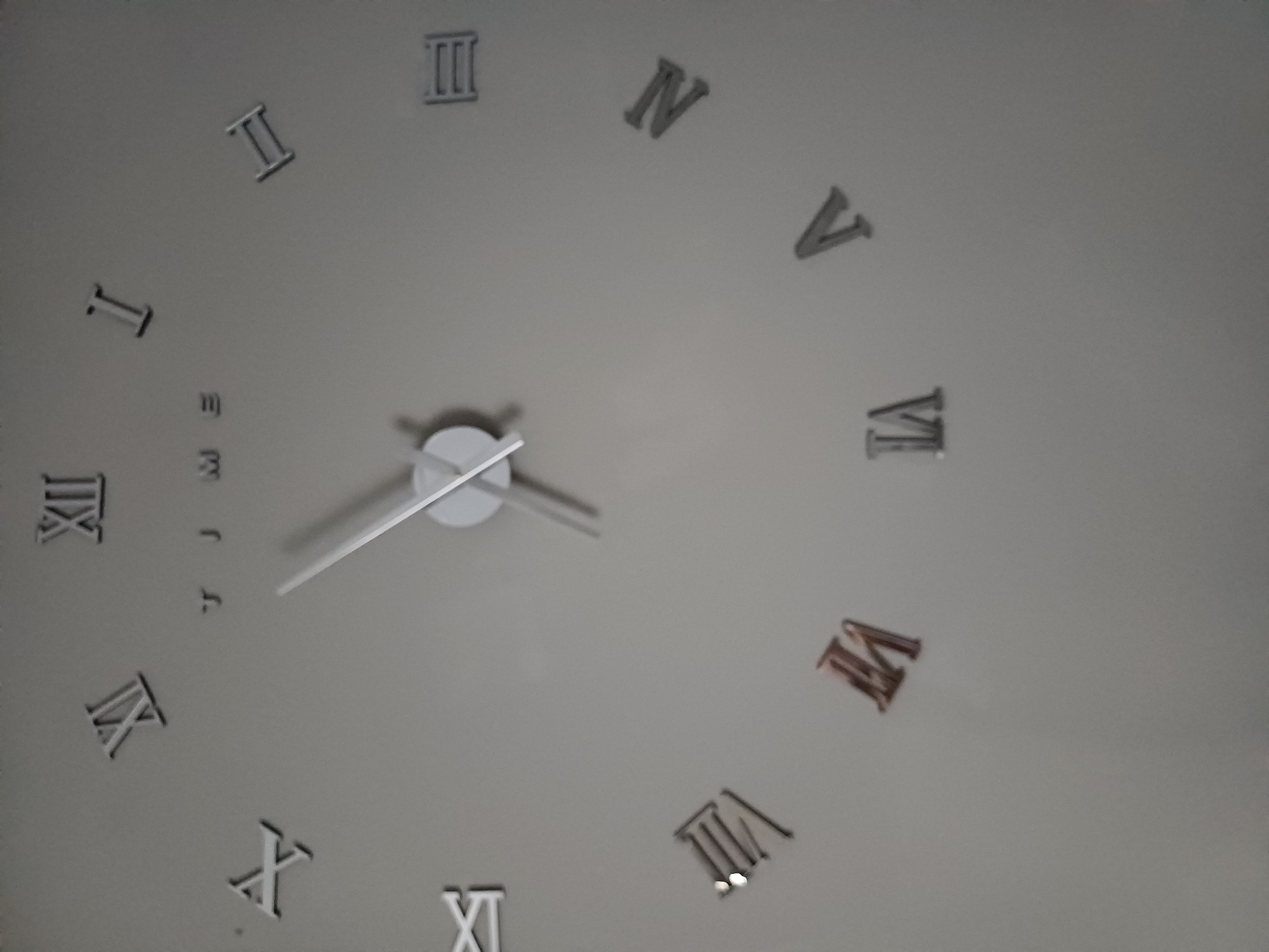 This is a photograph of a large, modern wall clock with Roman numerals, mounted on a plain gray wall. The clock appears to be a DIY or 3D type, with the numerals and hands being separate, adhesive pieces.
The Roman numerals visible are:
- XII at the top.
- XI to the left of XII.
- X to the left of XI.
- IV at the bottom left.
- V to the right of IV.
- VI to the right of V.
- VII to the right of VI.
- VIII to the left of VII.
- IX to the left of VIII.
- I to the right of XII.
- II to the right of I.
- III at the bottom right.
The clock hands are white and are positioned around the center. The hour hand is pointing roughly towards the IX position, and the minute hand is pointing roughly towards the III position. There is also text in the center of the clock, partially obscured by the hands, which appears to read "TIME".
The lighting in the image is somewhat dim, creating soft shadows around the clock elements, giving them a slightly raised, three-dimensional appearance. The overall background is a flat, neutral gray.
There are no people, animals, or other objects present in the image. The scene is static and focuses solely on the clock.
The question about the location in Sulymivka, Ukraine, is not supported by the visual information in the image. The image itself does not contain any clues or context that would place it in a specific city or country, including Sulymivka, Ukraine. Therefore, this detail cannot be provided based on the visual evidence.