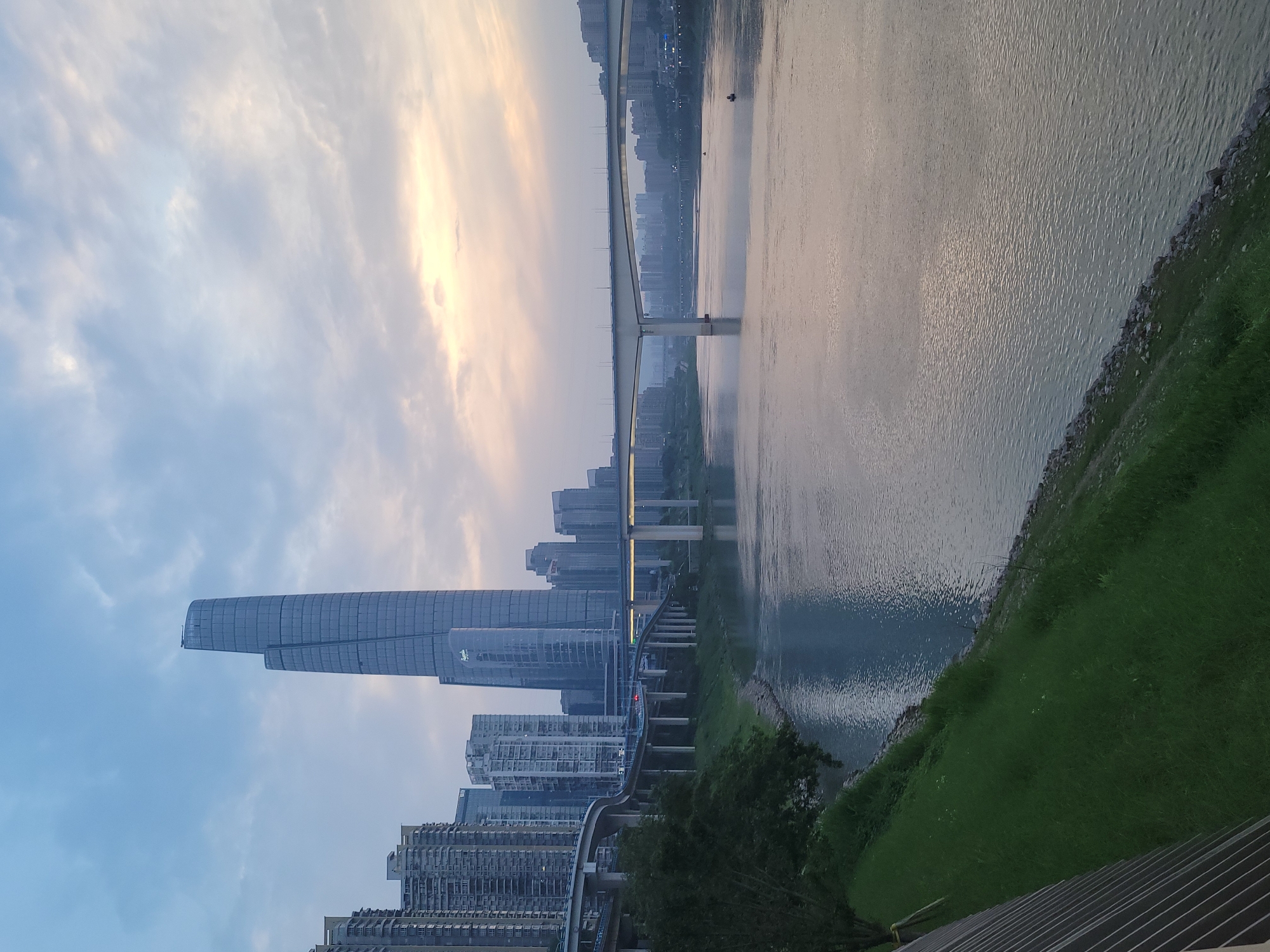 The image depicts a cityscape in Yuzhong District, China, at dusk or dawn. Dominating the view is a tall, modern skyscraper with a rounded, curved top, part of a cluster of high-rise buildings along the riverfront. A large river, likely the Yangtze, flows horizontally through the frame, its surface reflecting the light from the sky. A bridge spans the river, connecting different parts of the city. A light rail track or highway runs along the riverside, parallel to the river. The sky is a mix of blue and orange-yellow hues. Green vegetation is visible along the riverbank. There is a small boat in the water near the bridge. No people are directly visible in the image.