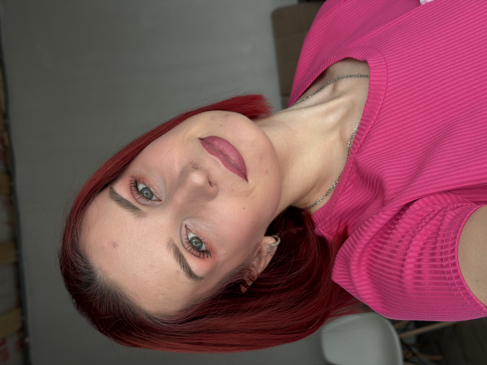 A woman with auburn hair is pictured indoors. She has fair skin, light eyes, and wears makeup. The lipstick and eyeshadow complement the bright pink top she is wearing. A silver chain necklace is visible around her neck. The background appears to be a room with neutral tones. The location is Chaikovsky, Russia.