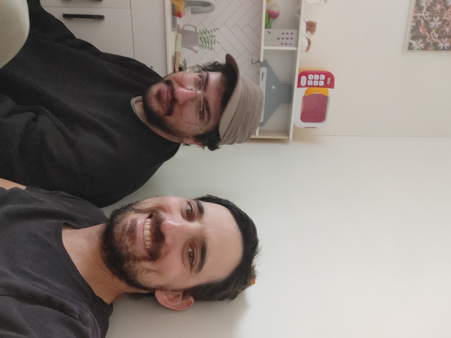 Two men are in the frame, seemingly indoors. The man in the foreground is wearing a dark t-shirt and has dark hair and a beard. He is smiling. The man behind him is wearing a black sweater, round glasses, and a baseball cap. Behind them is a white wall with a cabinet and some decorative items, including a toy. The location is Unknown, Unknown.