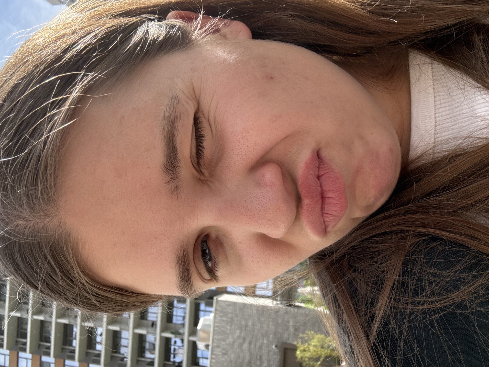 A close-up photo depicts a woman with brown hair and light skin. Her face is contorted with one eye closed and her lips pursed. In the background are buildings, indicating an urban setting. The location is Belgrade, Serbia.