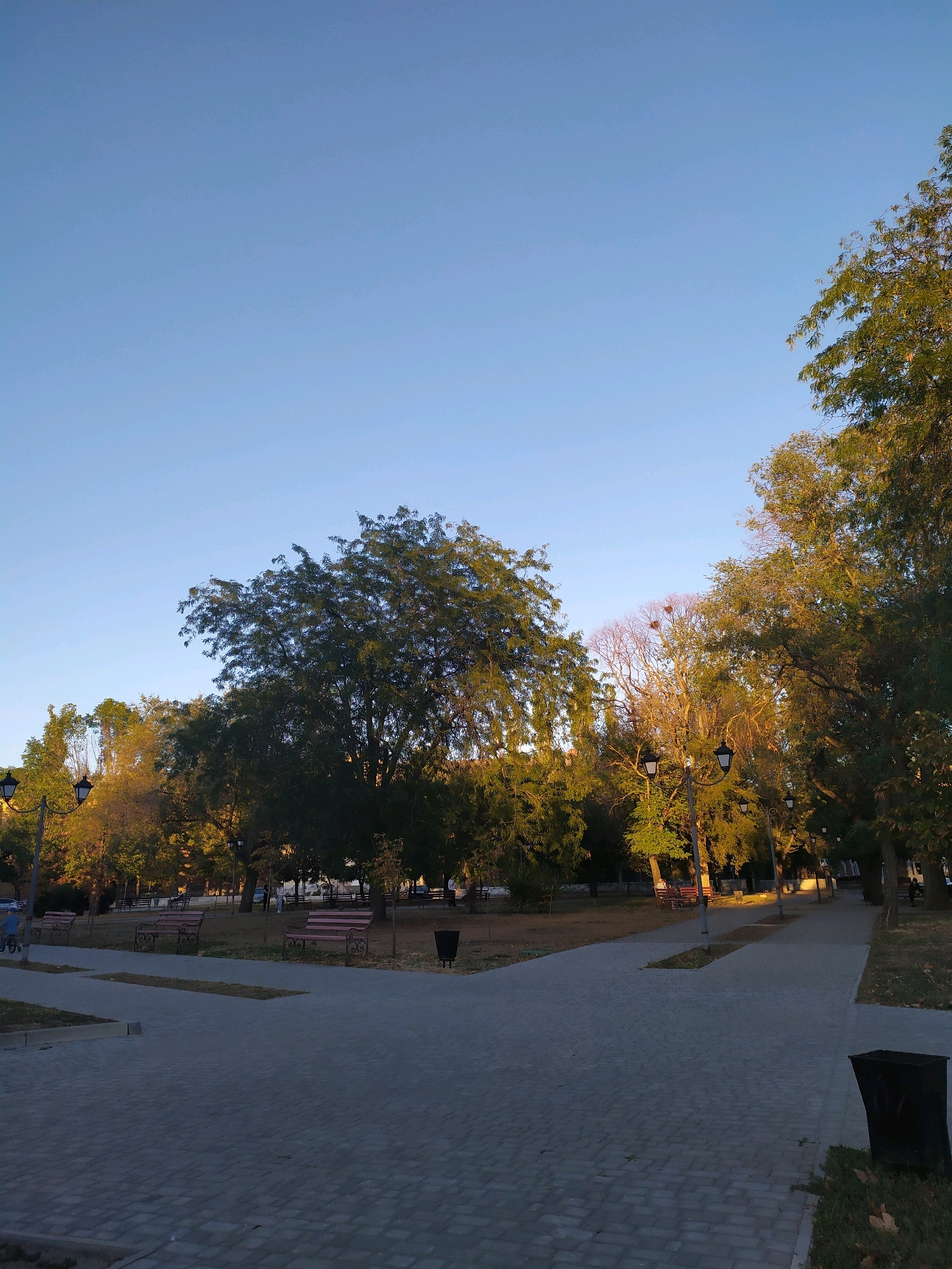 A view of a park in Mykolaiv, Ukraine, on a clear day. The sky is a pale blue, with the sun casting long shadows across the paved walkways and grassy areas. Several park benches are visible, some empty and others occupied by people. Trees with green foliage line the paths, with some showing autumnal colors. Lampposts are strategically placed throughout the park, illuminating the walkways. A trash can is visible in the foreground. The park appears to be a well-maintained public space, offering a tranquil setting for recreation.