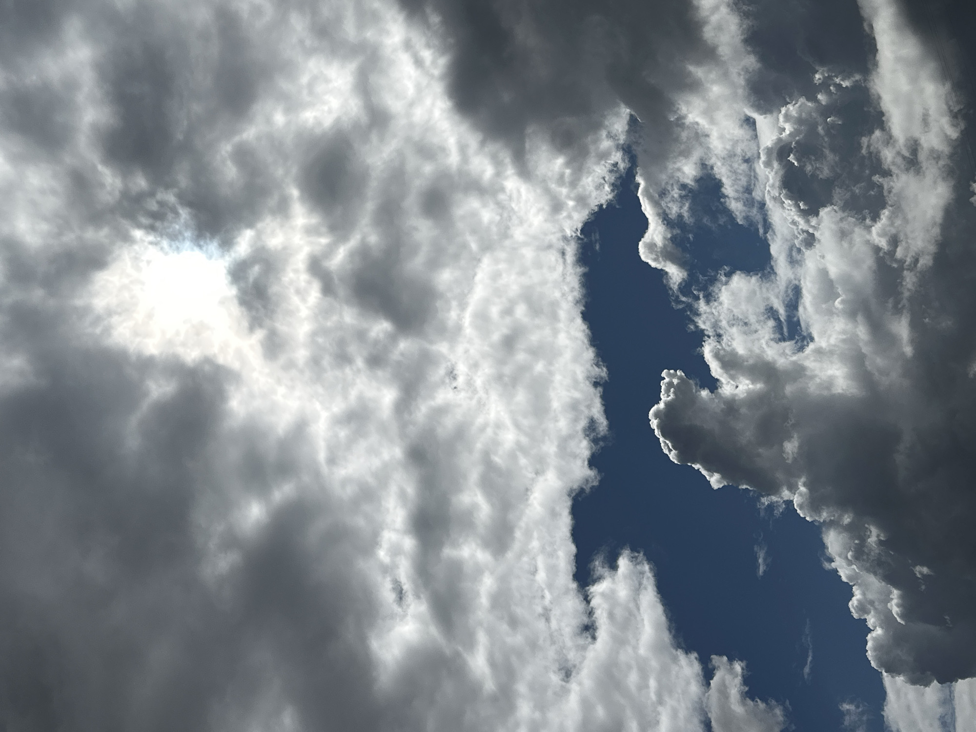 The image displays a cloudy sky over Podolsk, Russia. The clouds are varied, with some appearing dark and heavy, suggesting impending rain or a storm, while others are lighter and show hints of sunlight breaking through. The sunlight is partially obscured by the clouds, creating a dramatic contrast between light and shadow. The sky itself is a deep blue in the clearer patches. There are no people or man-made objects clearly visible in the frame. The scene is a natural landscape focused on atmospheric conditions.