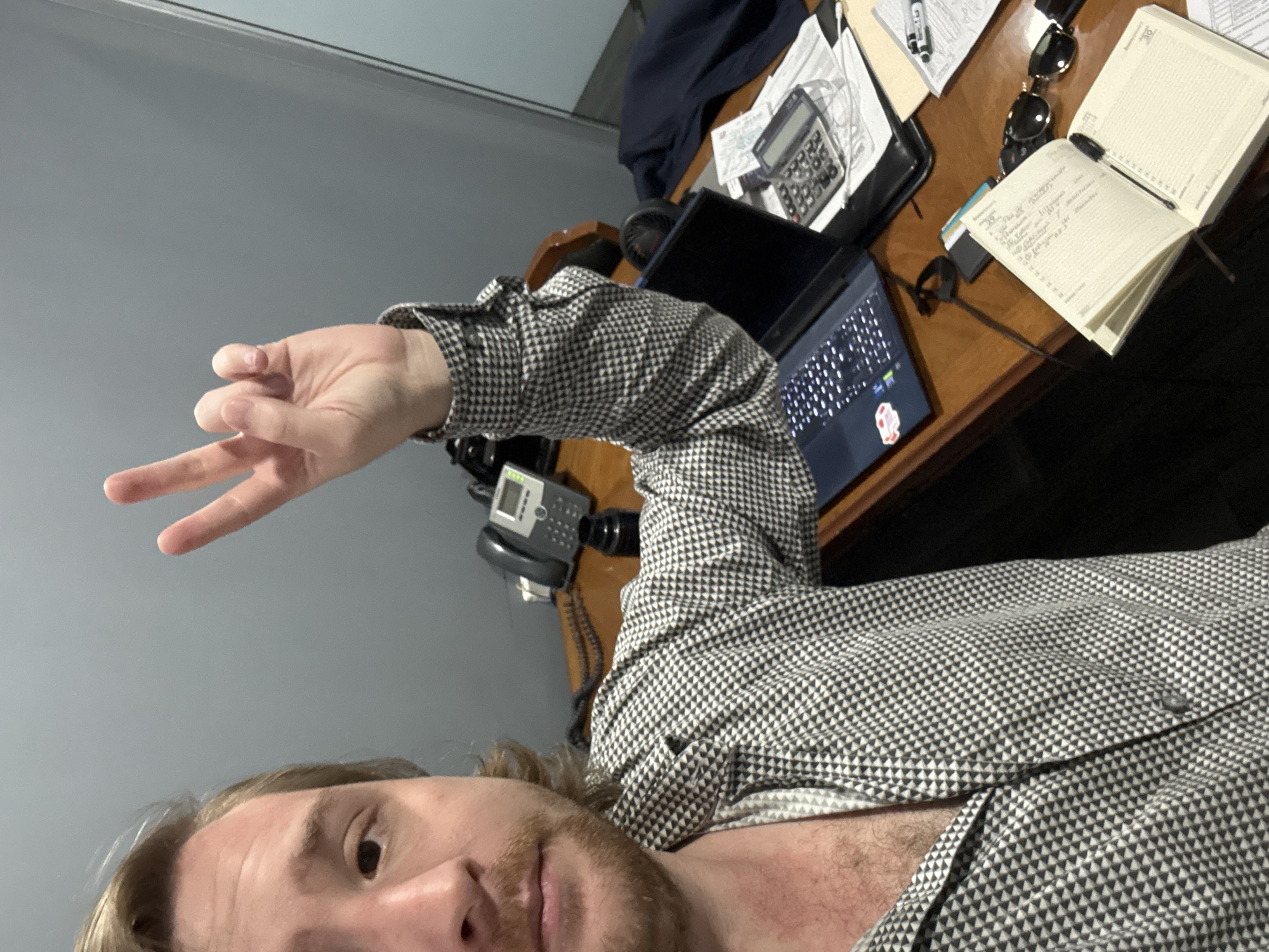 The image is a selfie taken indoors. The person in the image is fair-skinned with blonde hair and a beard. They are wearing a patterned button-down shirt and holding up two fingers in a "peace" sign. The background consists of a desk with various items including a laptop, a calculator, sunglasses, a notepad, and a phone. There is also a wall in the background. There is no information to suggest that this image was taken in Cuautitlán Izcalli, Mexico.