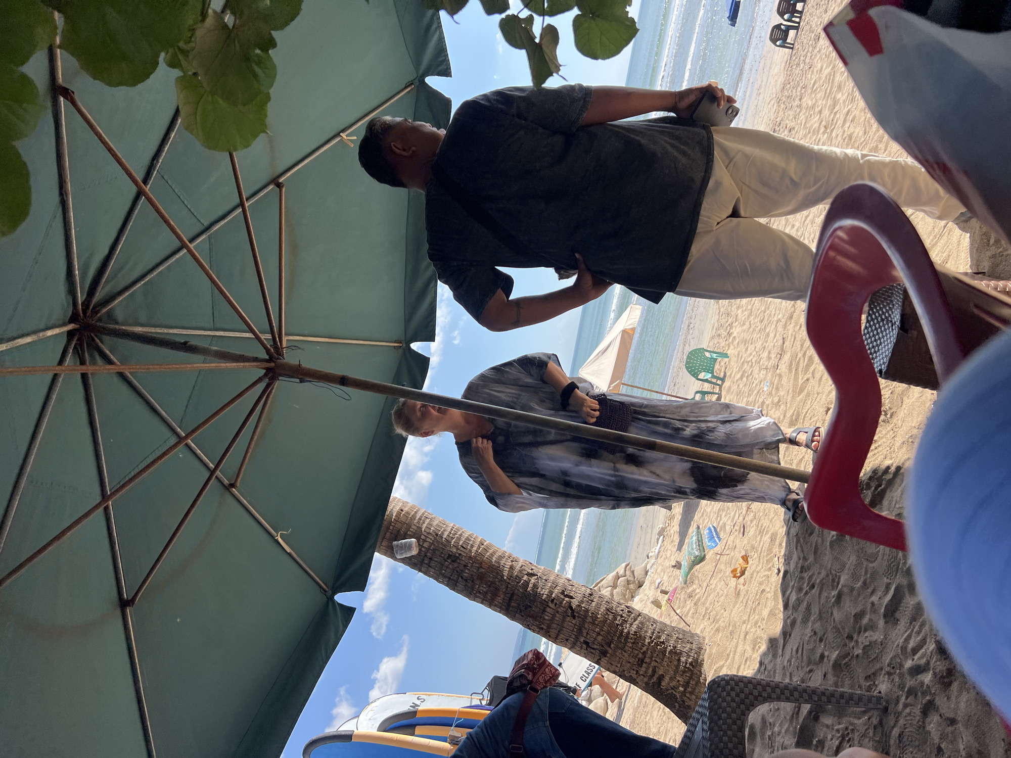 This image captures a beach scene in Kuta, Indonesia, under a large green umbrella. In the foreground, out of focus, are parts of red and blue chairs and what appears to be a person's head. Behind these, a woman stands facing the camera, wearing a grey and white tie-dye dress and holding a small bag. To her right, a man is seen from the back, wearing a dark t-shirt and light-colored pants, with one hand on his hip and the other holding a phone or small device. A palm tree is positioned between the woman and the ocean. The beach has sandy terrain, with several beach chairs and umbrellas visible in the background. The ocean and sky are also visible, with waves lapping at the shore and scattered clouds in the sky. A few surfboards are leaning against the palm tree.