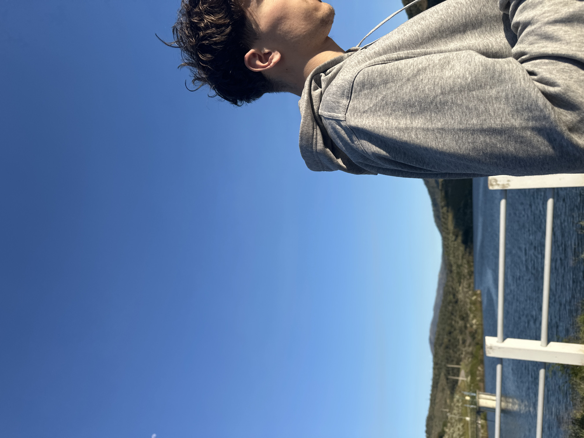 The image shows a man wearing a gray hoodie from the side, with his face tilted upwards toward a clear blue sky. He has dark, curly hair. The background consists of a body of water (likely a lake or river), a forested hillside, and a white railing in the foreground.
Without more information, pinpointing the exact location in Mirandela, Portugal, is not possible, but it's likely a spot near the Tua River or one of the nearby reservoirs, where there are overlooks with railings and views of the water and surrounding hills.