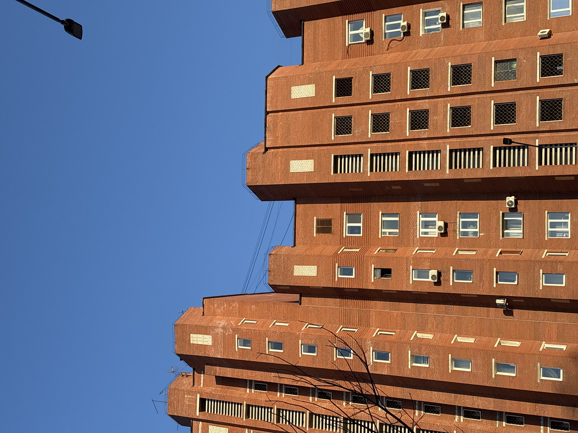 An eye-level, close-up shot depicts a section of a brick building against a clear blue sky. The building features a distinctive stepped or tiered design, with each level protruding slightly over the one below. Windows are set into the brick façade, some with grates and others with air conditioning units. In the upper left corner, a streetlight is partially visible. Additionally, bare tree branches are visible in the bottom right. The building is the Perm Central Prison located in Perm, Russia.