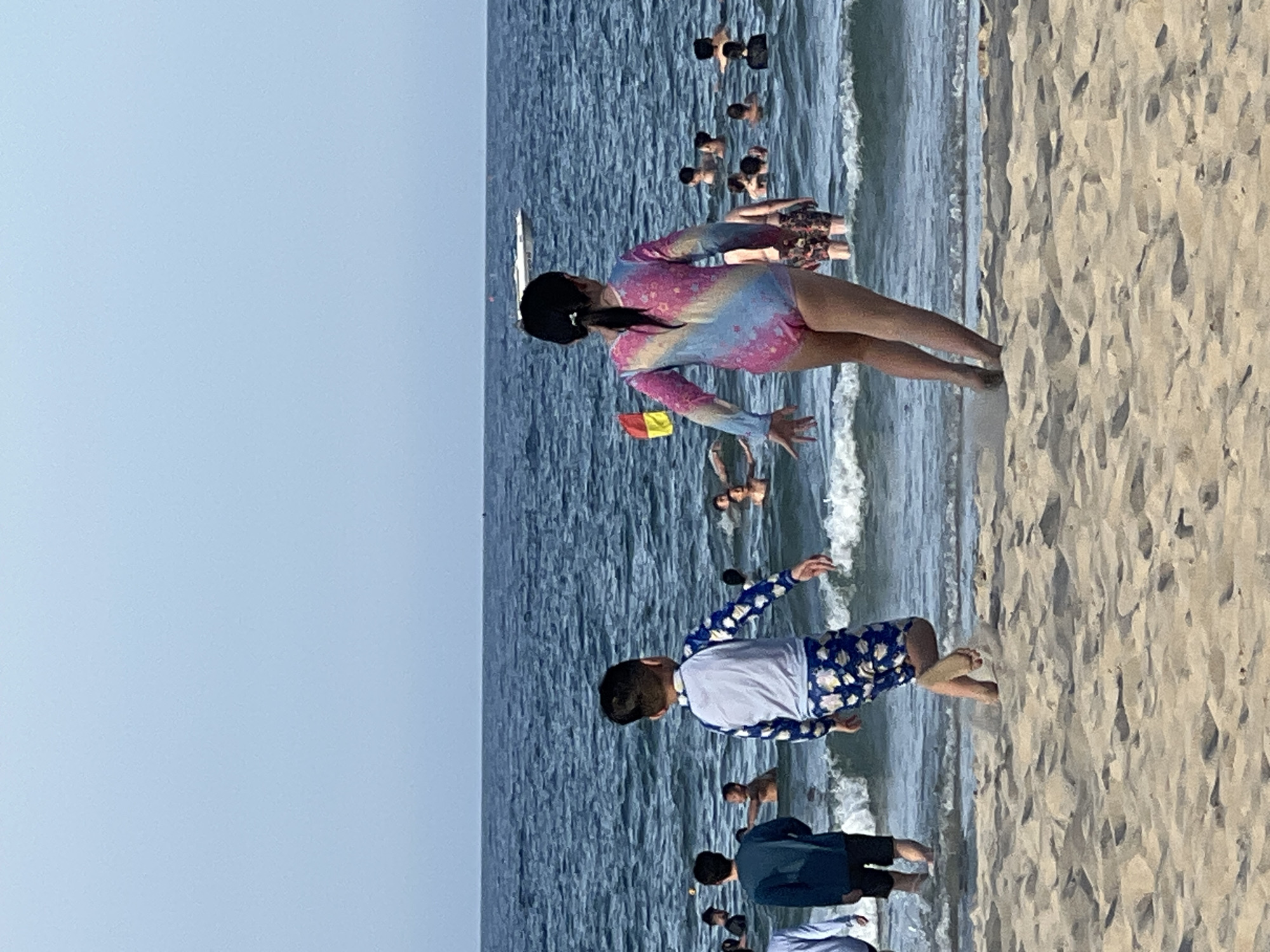 The image depicts a beach scene at Mỹ Khê Beach in Đà Nẵng, Vietnam. In the foreground, two children, a boy and a girl, are running towards the ocean from the sandy shore. The boy, on the left, wears a blue and white long-sleeved rash guard with a floral pattern on the shorts. The girl, on the right, is in a multi-colored one-piece swimsuit. In the background, numerous people are in the ocean, swimming and enjoying the water. The ocean is blue with moderate waves, and the sky is clear and light blue. A small white boat is visible in the distance on the water, and a yellow and red flag is planted near the shore. The sand is light brown and textured. The overall scene captures a typical summer day at a popular beach destination.
