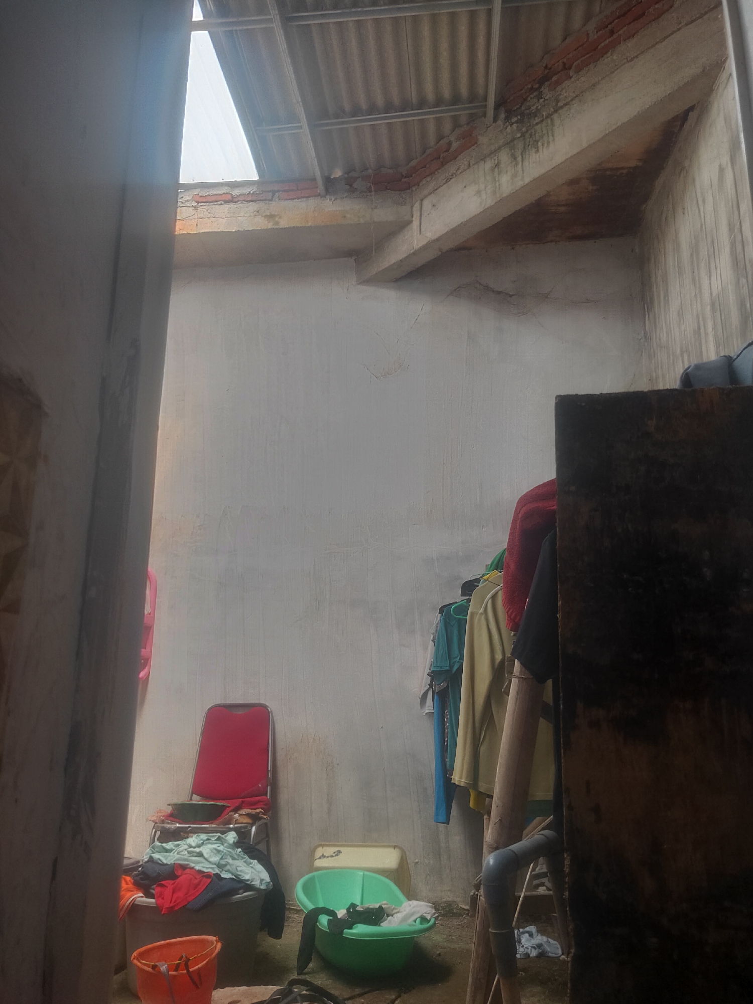 This image depicts an outdoor laundry area or a makeshift outdoor storage space in Antirogo, Indonesia. The scene is somewhat cluttered and appears to be utilitarian. In the foreground, a large, dark, rectangular object, possibly a piece of furniture or storage, dominates the right side of the frame. To the left, an open doorway reveals part of another room or structure with a patterned wall.
The central area shows a red chair with a green basin on top, surrounded by various laundry items piled in a large gray container and an orange bucket. Beside this, a green wash basin is visible. A clothesline with several garments hangs on the left side of the frame, supported by a wooden structure that also has a gray pipe attached. The walls are unfinished concrete, and the roof above is corrugated metal with exposed beams and some brickwork. A bright opening in the roof allows natural light to enter the space, casting shadows. The ground appears to be dirt or concrete. There are no people visible in the image.