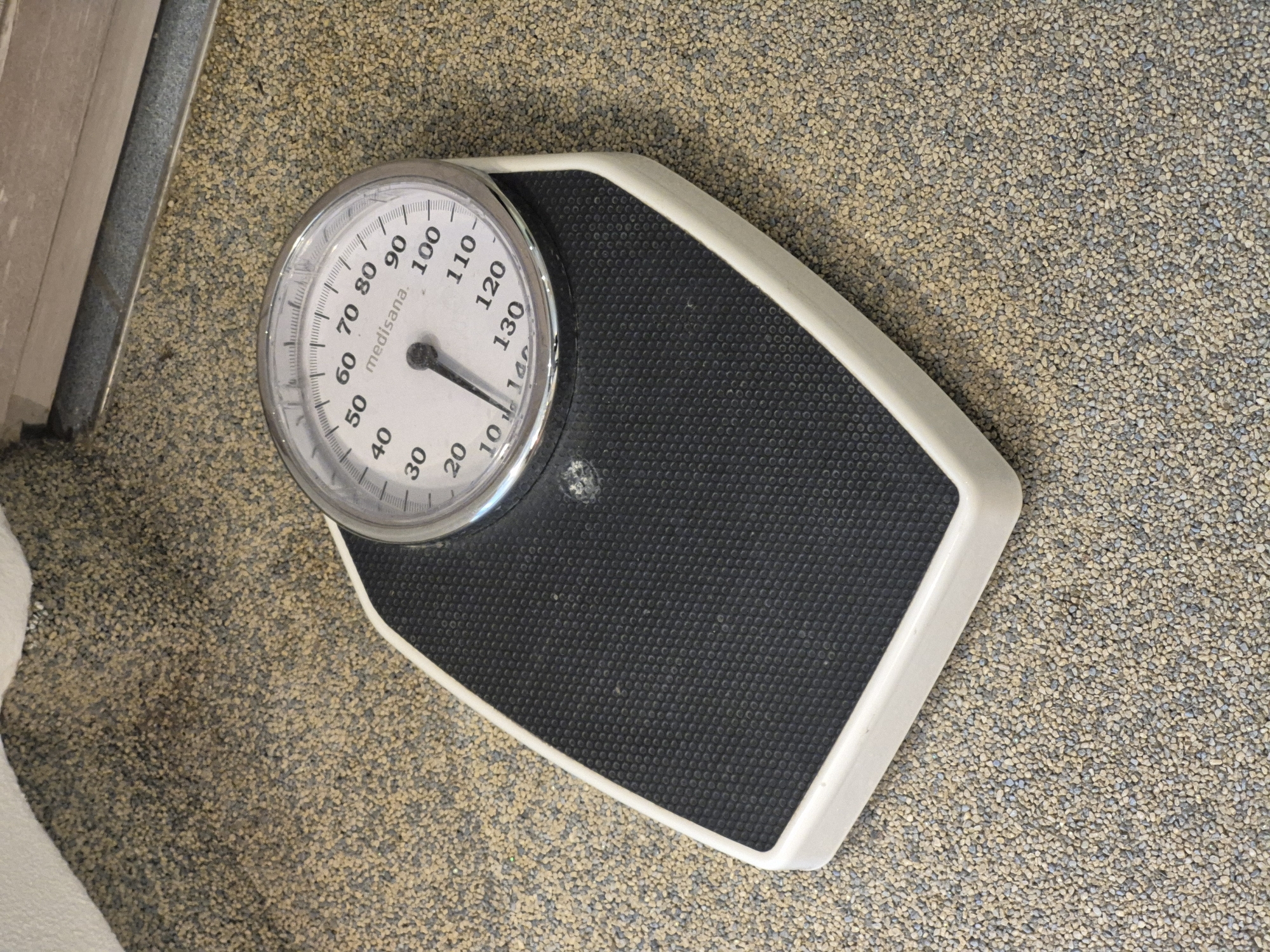A white and black analog bathroom scale is placed on a speckled beige and grey carpeted floor. The scale has a circular dial with a needle pointing slightly past the 40 mark. The dial displays numbers from 10 to 140, with markings every 10 units and smaller tick marks every 2 units. The brand name "medisana." is visible on the dial. The scale has a black textured surface where one would stand, and a white plastic rim. The background shows a portion of a wall with a metallic trim. The scene is indoors. The location is the city of Schwäbisch Gmünd, Germany.