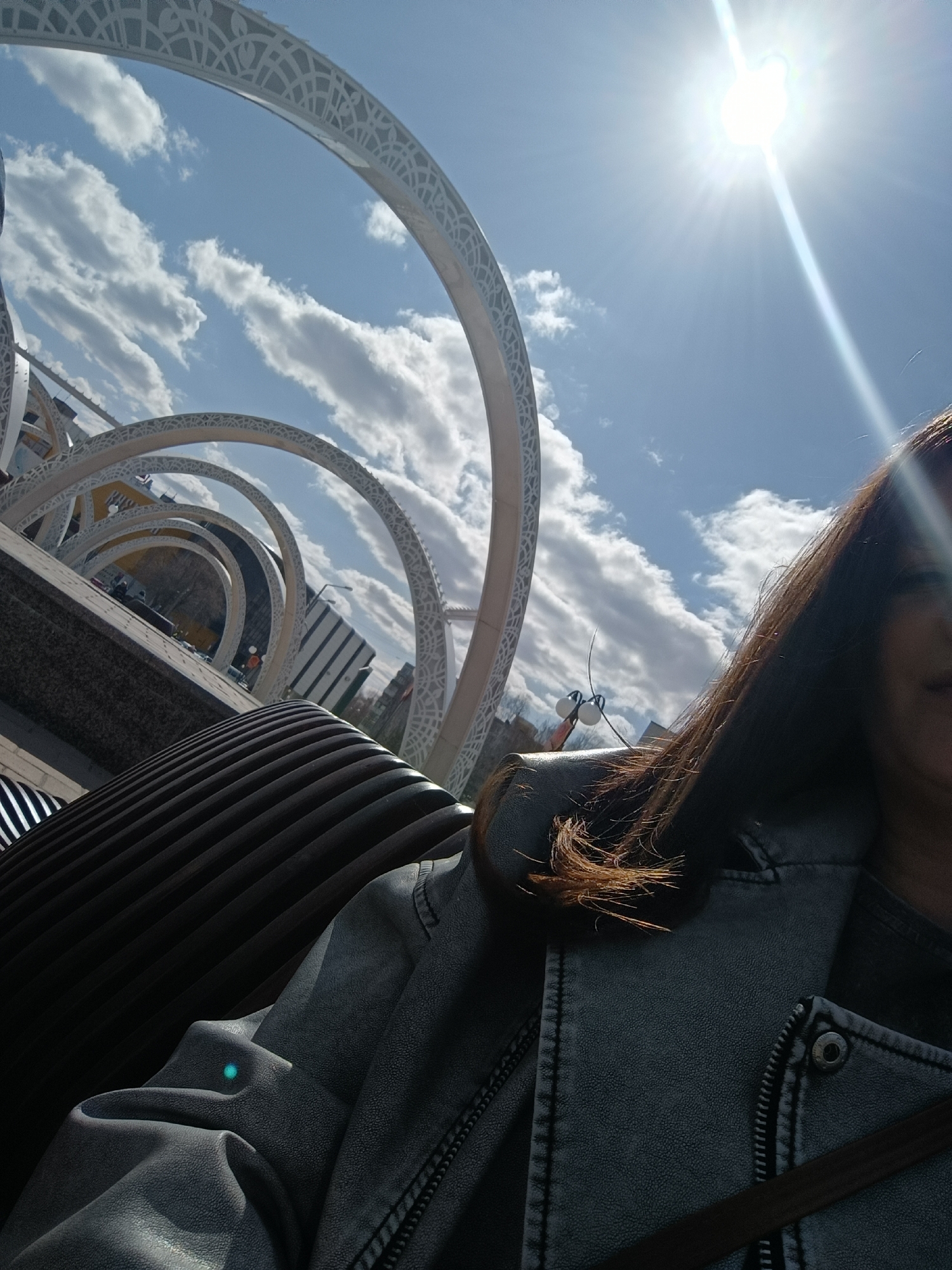 A person is sitting on a bench in front of an art installation in Tobolsk, Russia. The installation consists of a series of white, ornate arches that recede into the distance under a blue sky with scattered clouds. The sun is shining brightly overhead. The person is wearing a grey jacket and their dark hair is visible in the foreground. The bench is made of dark slats and has a metallic frame. Buildings can be seen in the background, partially obscured by the art installation.