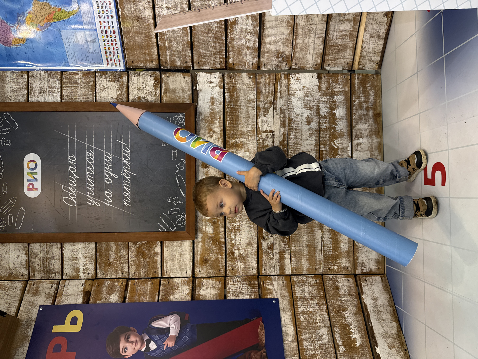 A young boy, appearing to be about 3-4 years old, stands in a room with a rustic wooden wall backdrop. He is holding a large, light blue pencil horizontally. The boy is wearing a dark gray sweatshirt, blue jeans, and sneakers. The pencil has the word "РИО" in rainbow letters on it. To the left of the boy is a large poster featuring a boy in a blue vest and bow tie, with the Cyrillic letters "рь" visible. To the right of the boy, a world map is displayed. A chalkboard with handwritten Russian text and drawings of school supplies is mounted on the wall behind the boy. A wooden ruler with markings is leaning against the wall to the far right. The floor is tiled and has a grid pattern, with a large red number "5" visible on the floor in the foreground. The lighting appears to be artificial, casting soft shadows. The overall scene suggests a themed play area or educational setting. The location is identified as Korobovo, Russia.