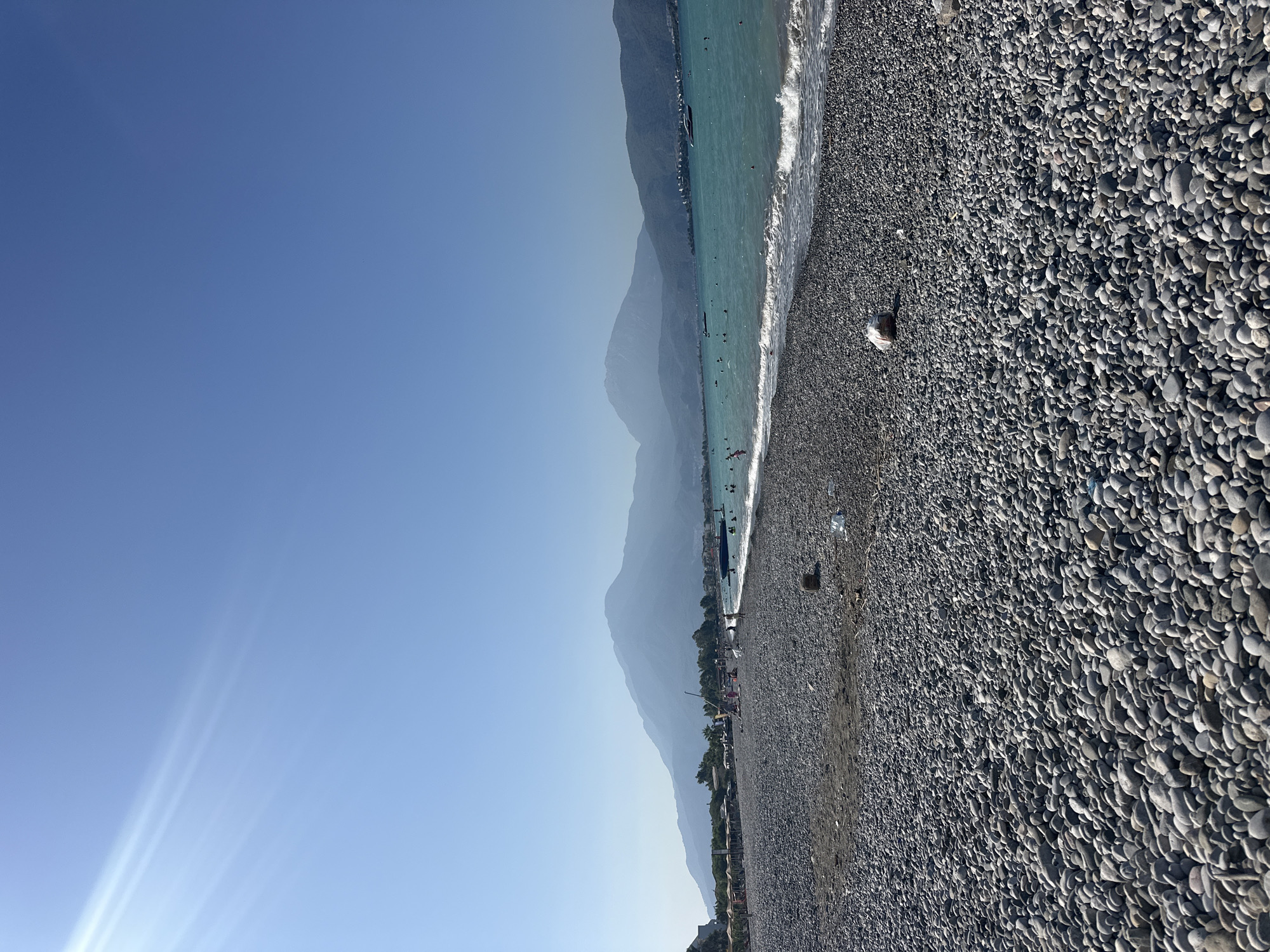 This image captures a scenic view of a pebble beach in Fethiye, Turkey, under a clear blue sky. The sun's rays are visible on the left side of the frame, casting a bright glow. The foreground is dominated by a vast expanse of smooth, grey pebbles, characteristic of many beaches in the region.
In the middle ground, the turquoise waters of the sea gently lap against the shore, with small waves creating white foam. Several boats are dotted on the water, and numerous people are visible swimming and enjoying the sea. Beyond the water, a lush green treeline lines the coast, giving way to imposing, hazy mountains in the background. The mountains, partially obscured by atmospheric conditions, form a majestic backdrop to the coastal scene. A few beach umbrellas and loungers are set up on the sand behind the pebble area, suggesting a developed tourist spot. The overall atmosphere is one of a peaceful, sunny day at the beach.