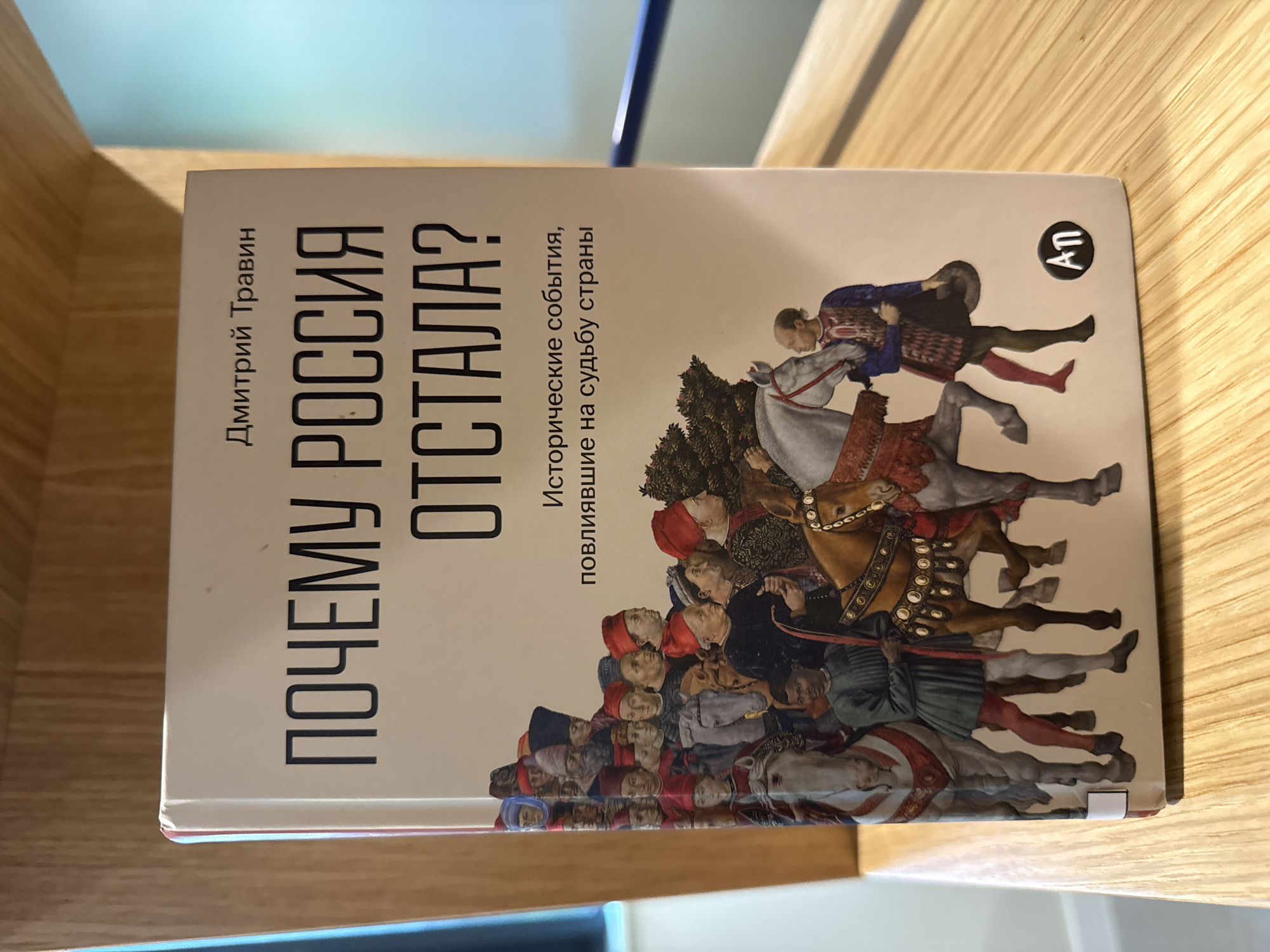 A hardcover book titled "Почему Россия отстала?" (Why did Russia fall behind?) by Дмитрий Травин (Dmitry Travin) is displayed on a wooden shelf. The book's cover features a painting of people in historical attire, many on horseback, in a procession. The background of the book cover is a light beige color. Below the main title, there is a subtitle: "Исторические события, повлиявшие на судьбу страны" (Historical events that influenced the fate of the country). In the bottom right corner of the book cover, there is a circular logo with the letters "АП" inside. The book is positioned on a light-colored wooden shelf with visible grain. The shelf is part of what appears to be furniture, with a blurred background suggesting an indoor environment. There is no indication or visual element in the image that places this scene in the city of Dilijan, Armenia.